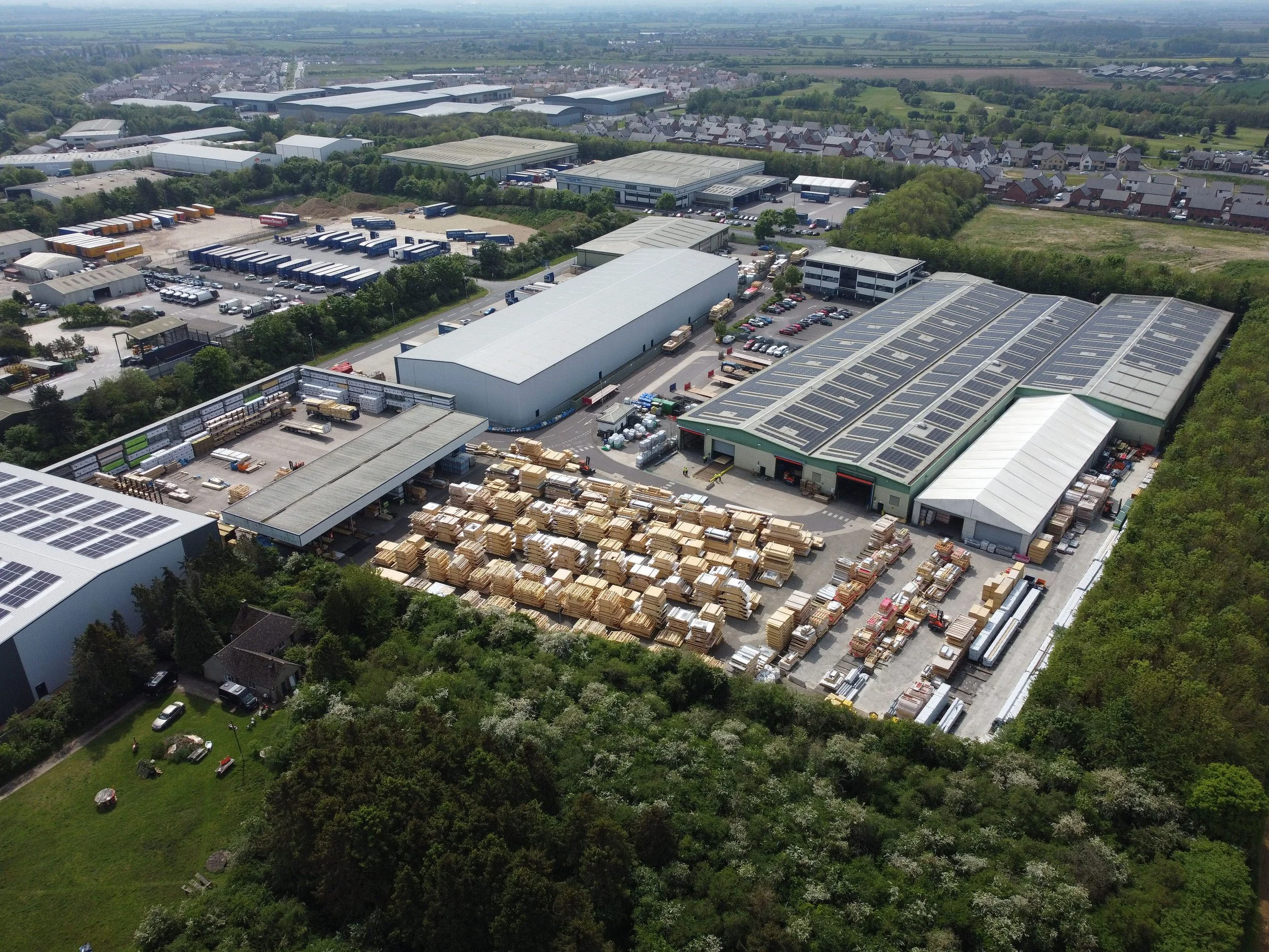 Aerial view of an industrial warehouse area with multiple buildings, solar panels on roofs, storage of wood and materials outside, surrounded by greenery and a residential neighborhood in the background.