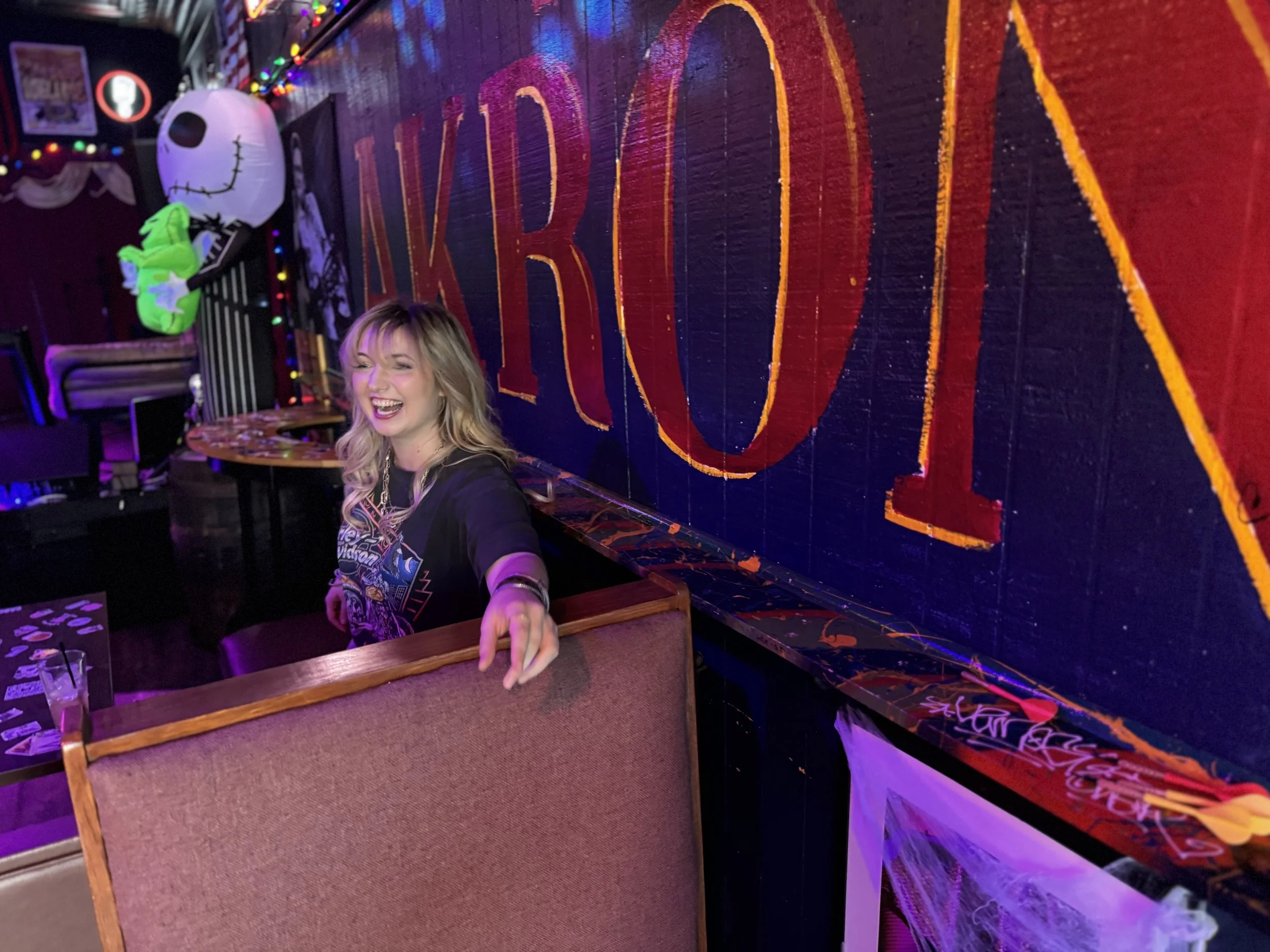 A young woman with blonde hair, smiling and pointing at the camera, sitting in a cozy, dimly lit place with Halloween decorations, including a large lantern resembling a skull and a green plush toy on a shelf. The background features a dark wall with