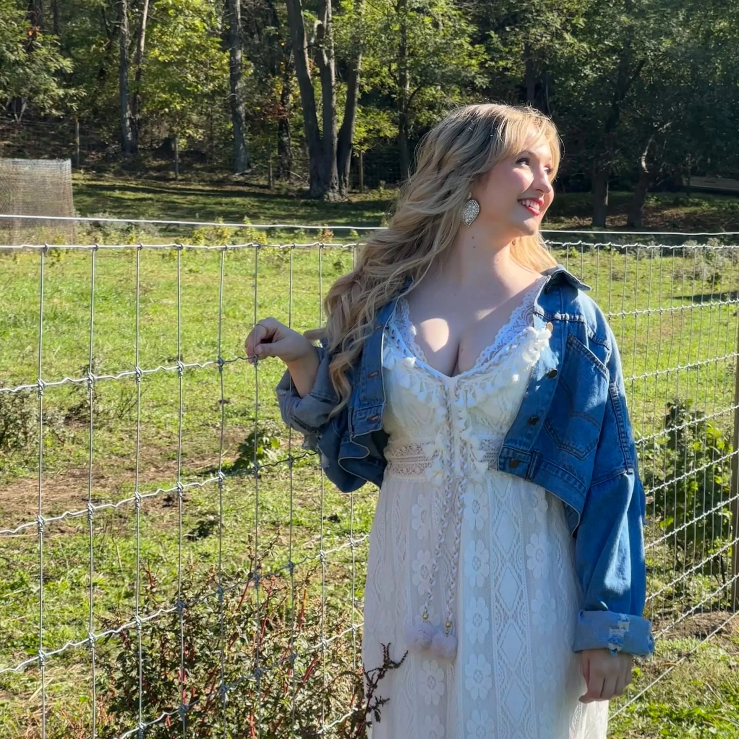 A woman with long blonde hair wearing earrings, a denim jacket, and a white lace dress standing outdoors next to a wire fence, smiling and looking to her right, with trees and grass in the background.