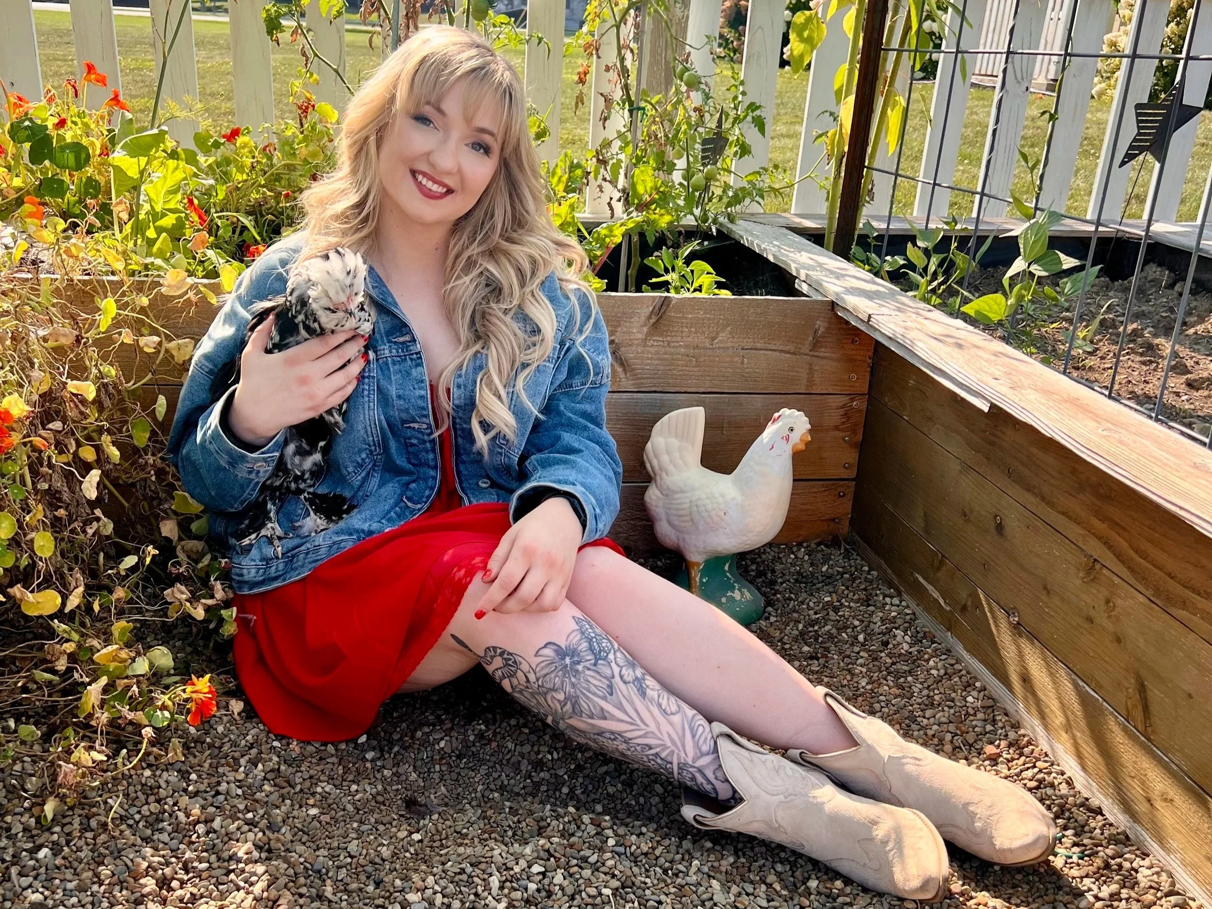 A young woman with blonde hair, wearing a denim jacket and red dress, sitting on gravel ground in a garden with plants and flowers, holding a black-and-white kitten, with a decorative chicken figure beside her.