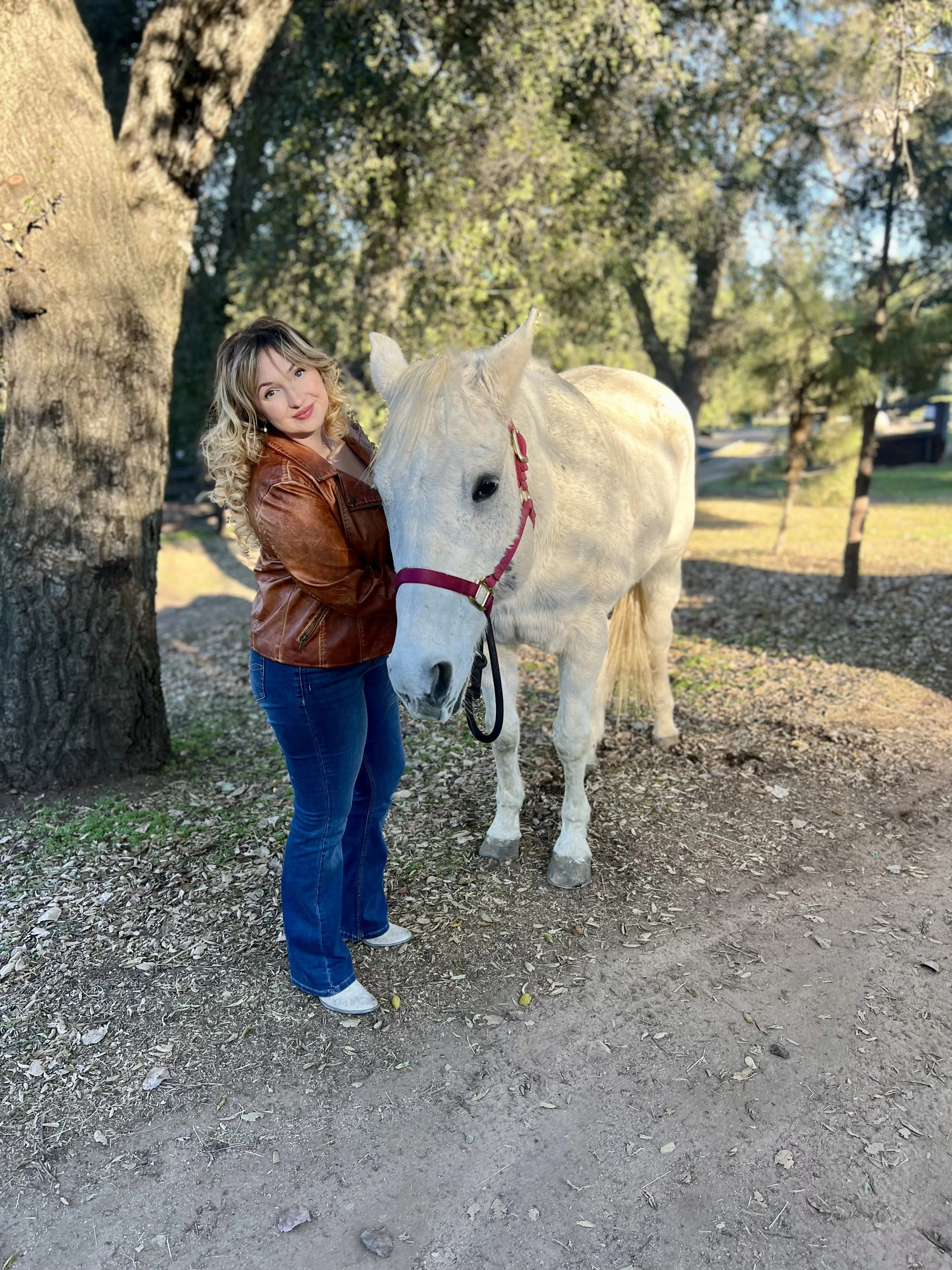 A woman with curly blonde hair wearing a brown leather jacket and blue jeans standing next to a white horse with a red halter, outdoors in a park with trees and sunlight.