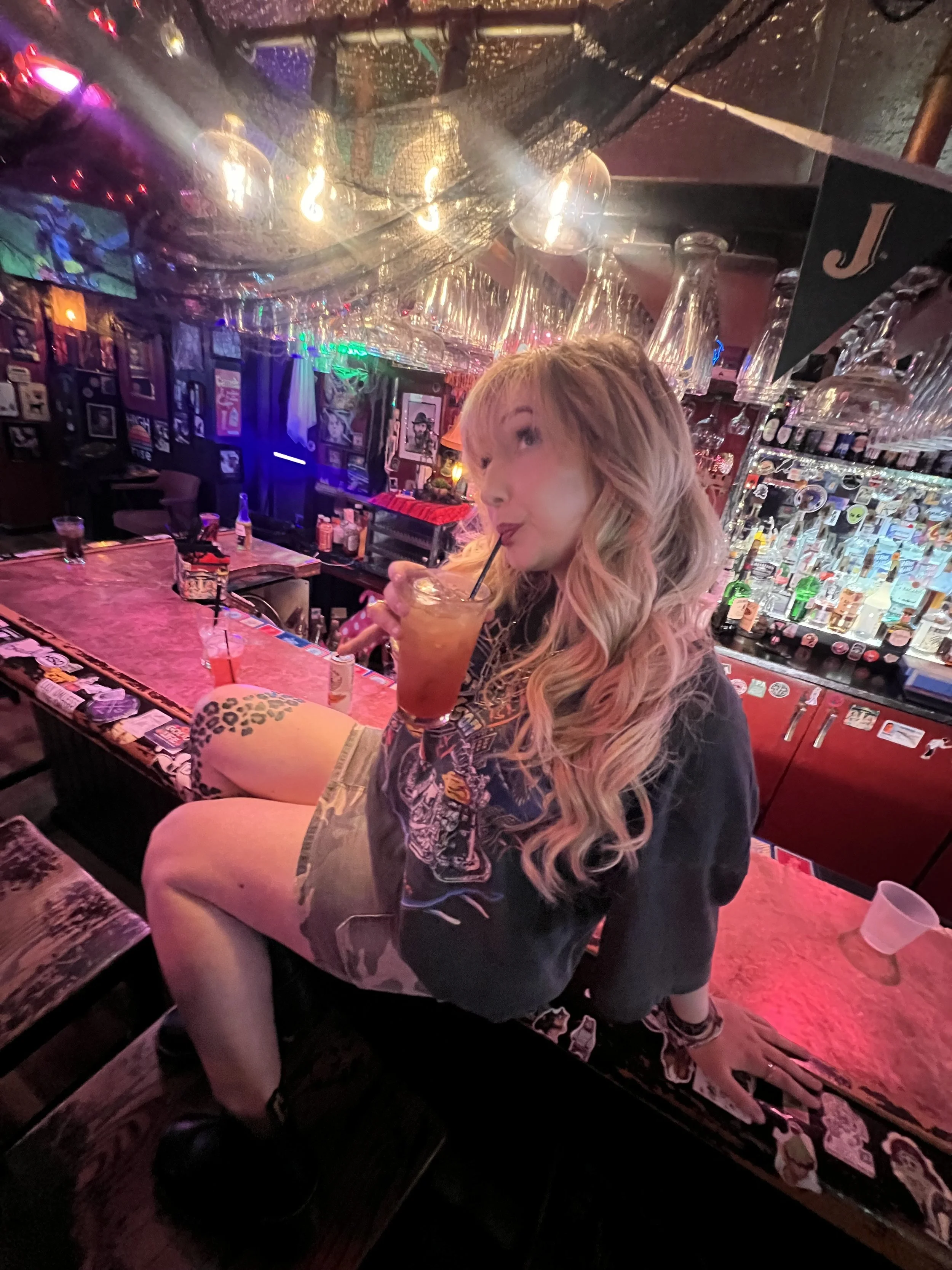 A young woman with long, wavy blonde hair sitting on a bar counter, sipping a drink through a straw, in a dimly lit bar with colorful neon lights, bottles, and glasses in the background.