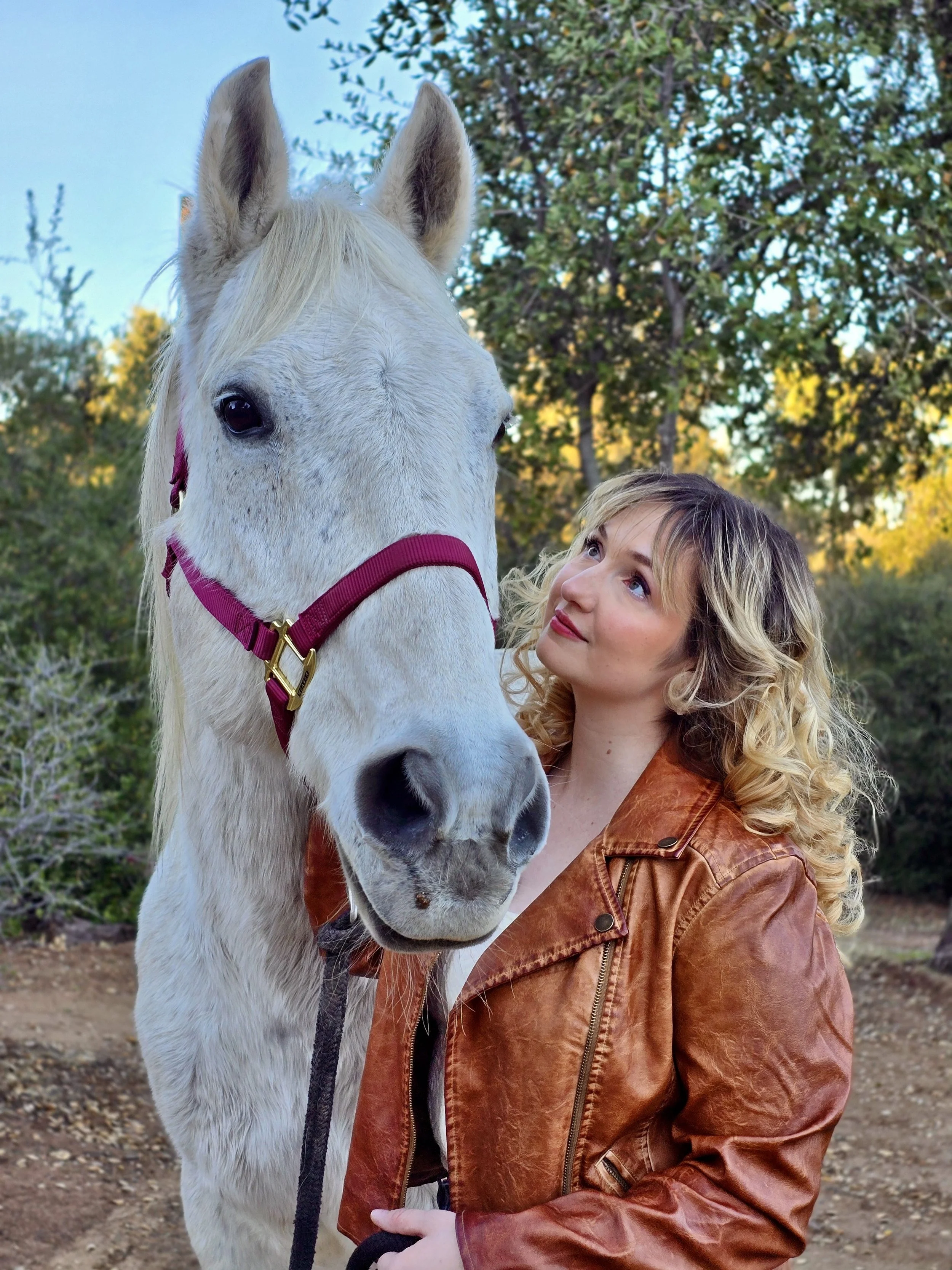 A woman with curly blonde hair wearing a brown leather jacket standing next to a white horse with a pink halter outdoors in a natural setting with trees and blue sky.