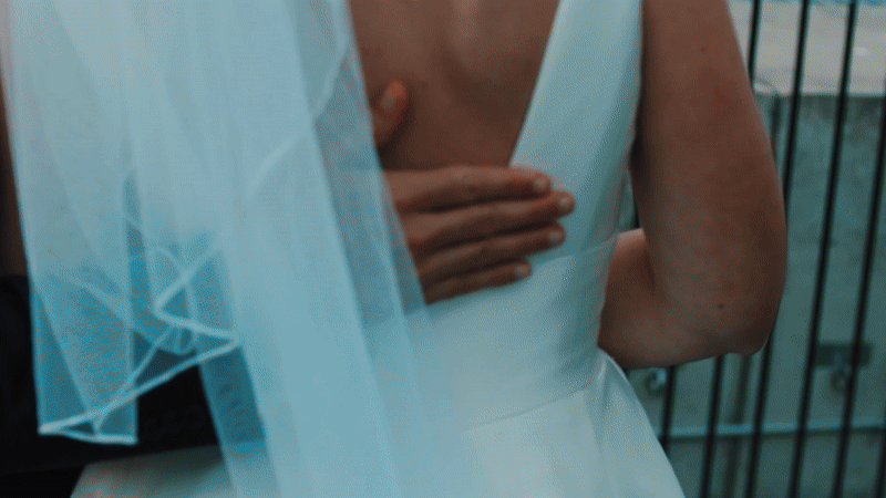 Close-up of a woman in a white dress with a sheer blue veil, standing on a balcony with black railings.
