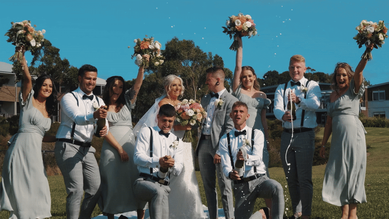 A group of ten people, including a bride and groom, celebrating outdoors with bouquets of flowers, some holding champagne bottles, and confetti falling around them on a sunny day.
