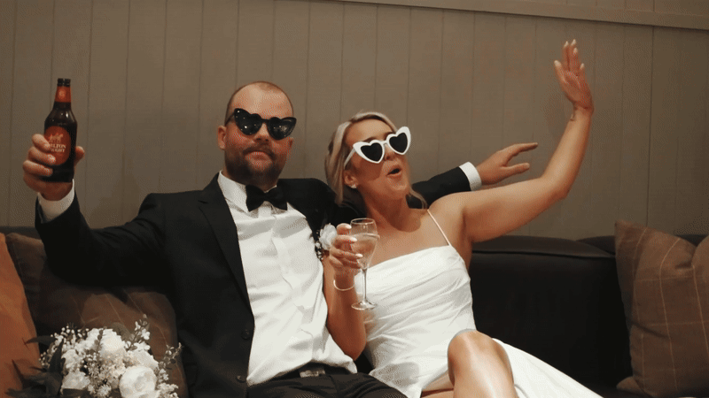 Man and woman dressed in formal attire wearing sunglasses, sitting on a couch, holding drinks, and celebrating.