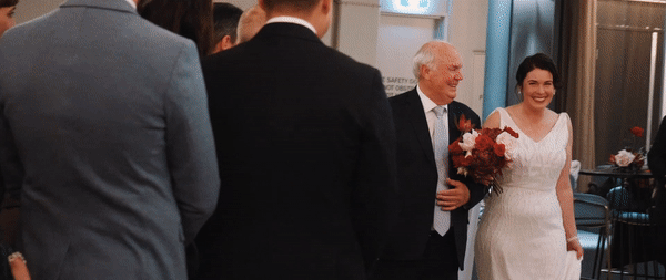 A bride in a white wedding dress holding a bouquet, smiling, walking down the aisle with an older man in a suit, at a wedding ceremony.