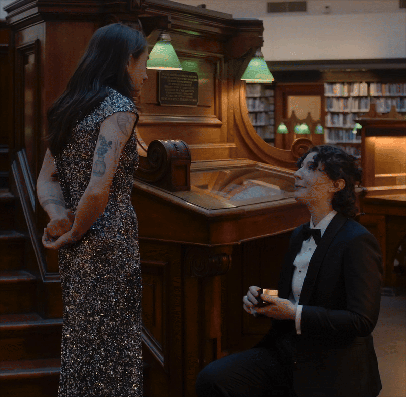A woman in a sparkly dress stands in front of a man in a tuxedo in a library with wooden furnishings and a vintage piano, engaging in a proposal.