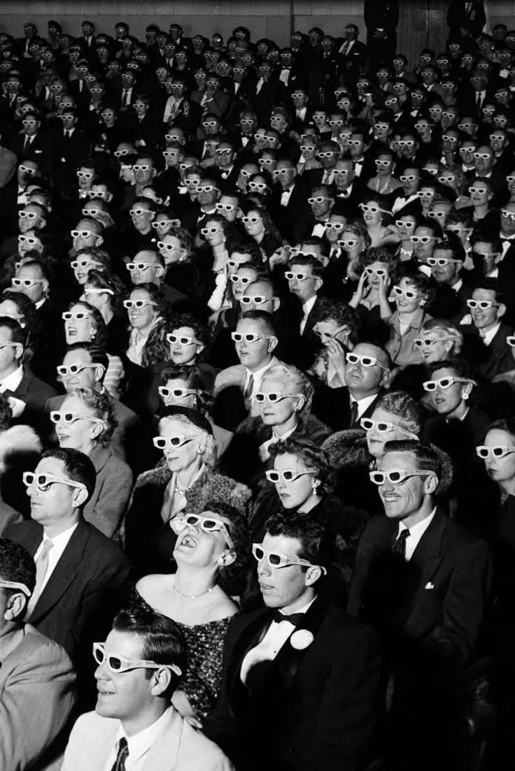 A large audience of men and women wearing 3D glasses attending a movie theater or presentation.