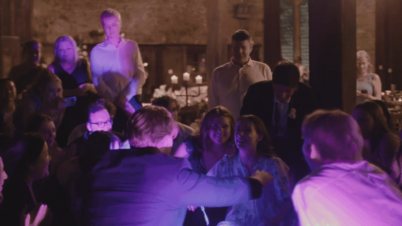 Group of people gathered around at a party, with some smiling and talking, in a dimly lit room with candles on the tables.