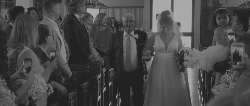 A bride walking down the aisle with her father in a wedding ceremony, surrounded by guests taking photos.