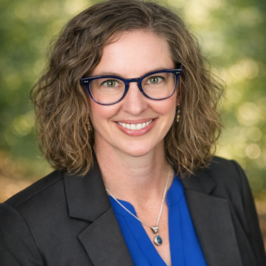 A woman with curly brown hair wearing glasses, a black blazer, and a blue blouse, smiling outdoors with a blurred green background.