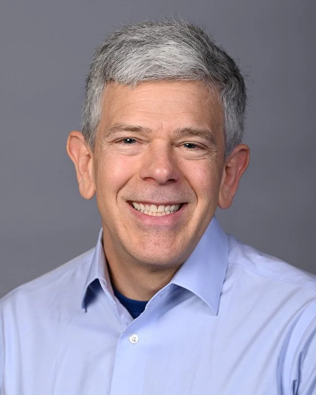 Smiling middle-aged man with short gray hair wearing a light blue collared shirt against a gray background.