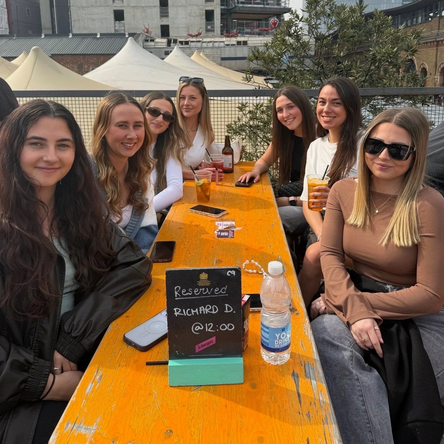 Group of seven women sitting at a long outdoor wooden table, smiling with drinks, a reserved sign for Richard D. at 12:00, and a water bottle.