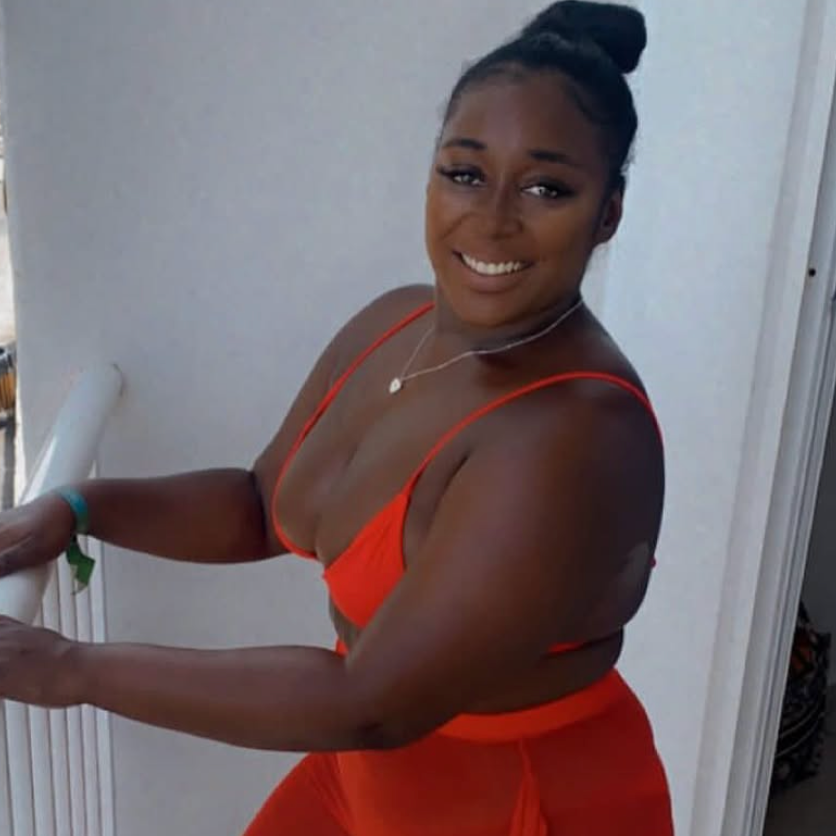 A smiling woman with dark hair in a bun wearing a red top and matching skirt, standing on a balcony with a white wall behind her.