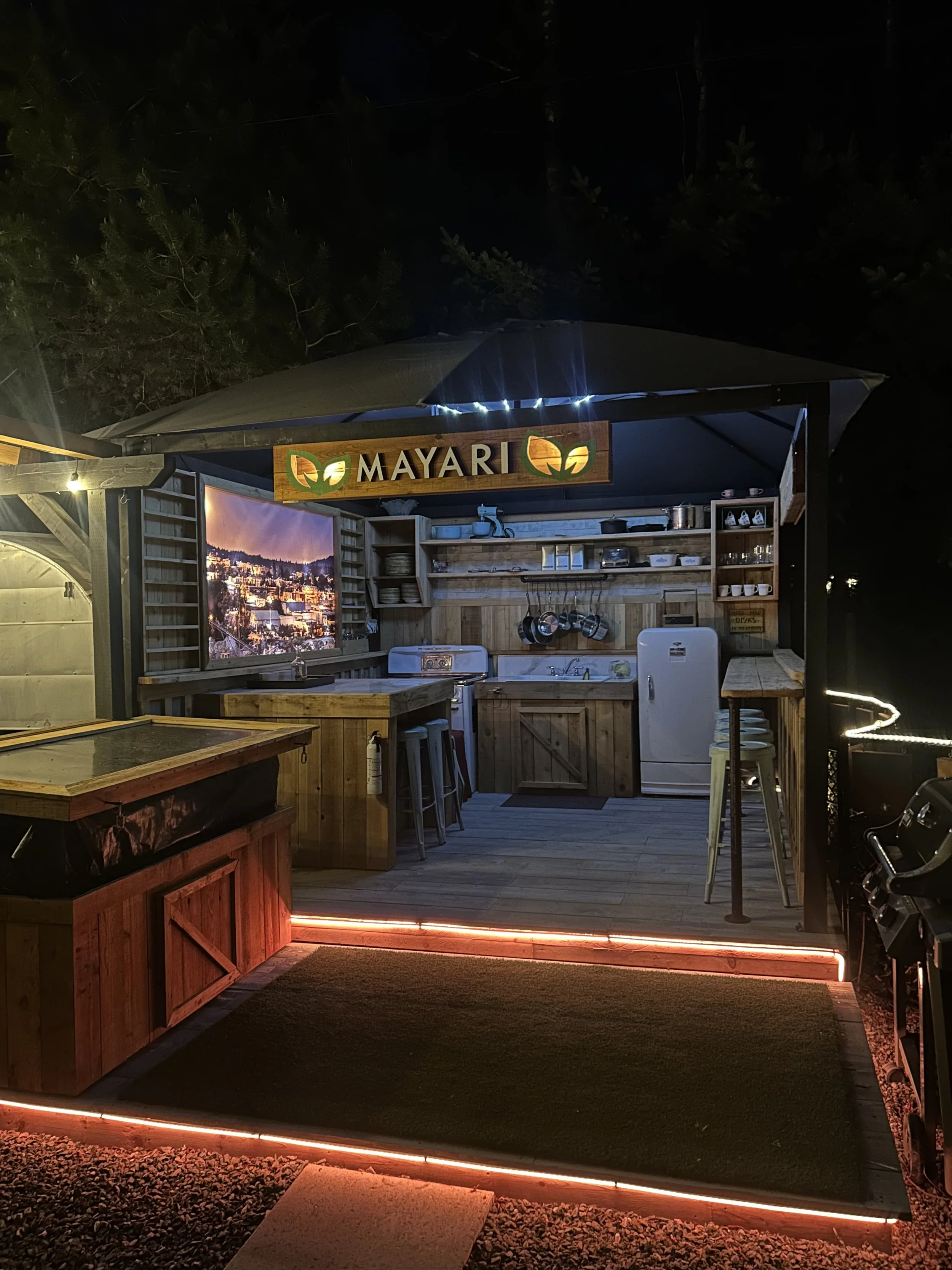 A small outdoor kitchen with a wooden sign that says 'MAYARI' with leaf designs, illuminated by string lights at night, featuring a sink, shelves with dishes, and a refrigerator, with a black rug in front and a sunset cityscape on the banner.