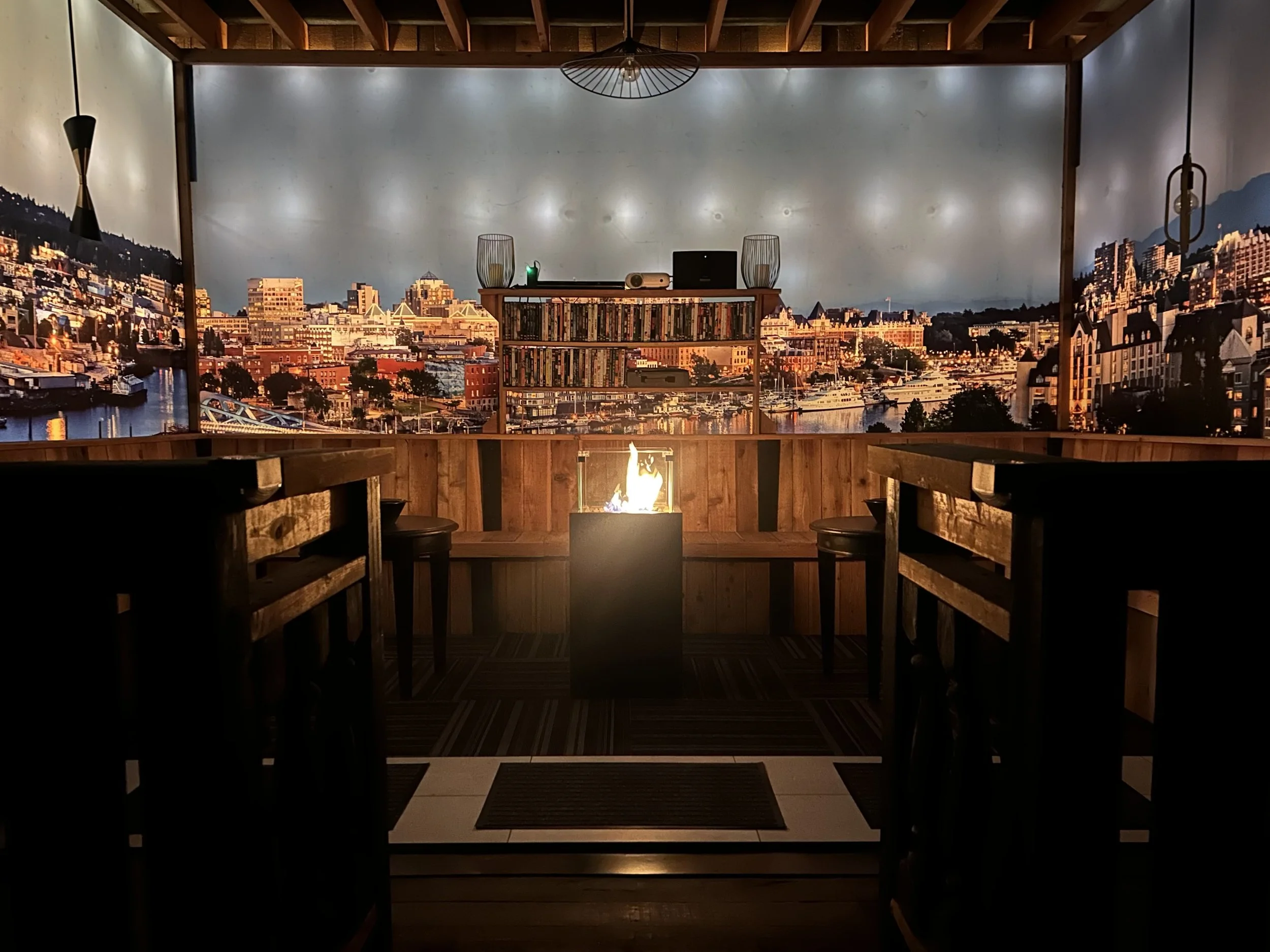 An interior view of a semi-enclosed balcony or porch with a cityscape mural on the wall, a small fire pit in the center, a bookshelf with books, and wooden bar stools around the fire pit.