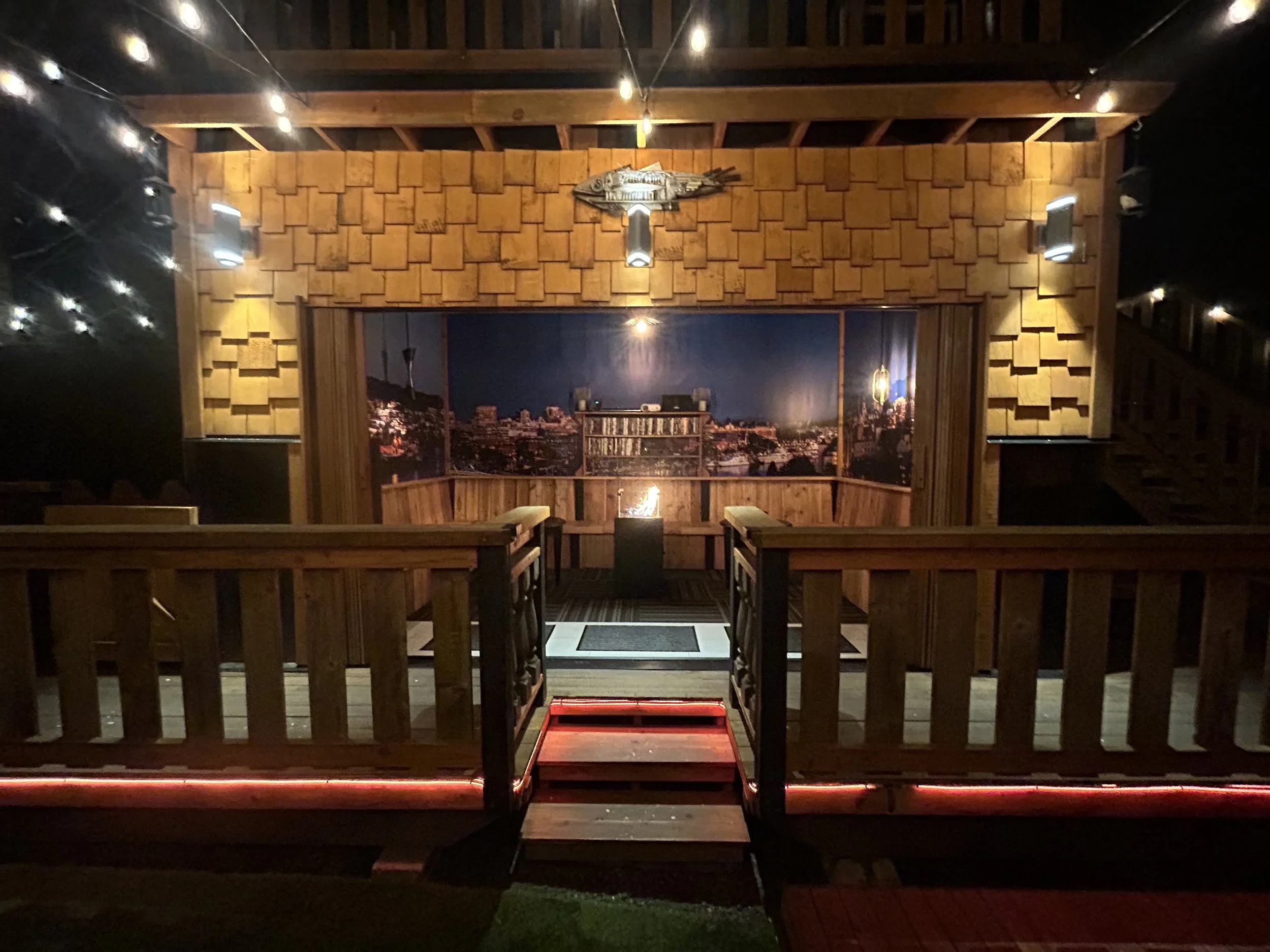 A cozy indoor space with wooden walls and warm lighting, featuring a scenic cityscape mural on the back wall. There are small tables and chairs, and string lights hanging from the ceiling, creating a warm ambiance.