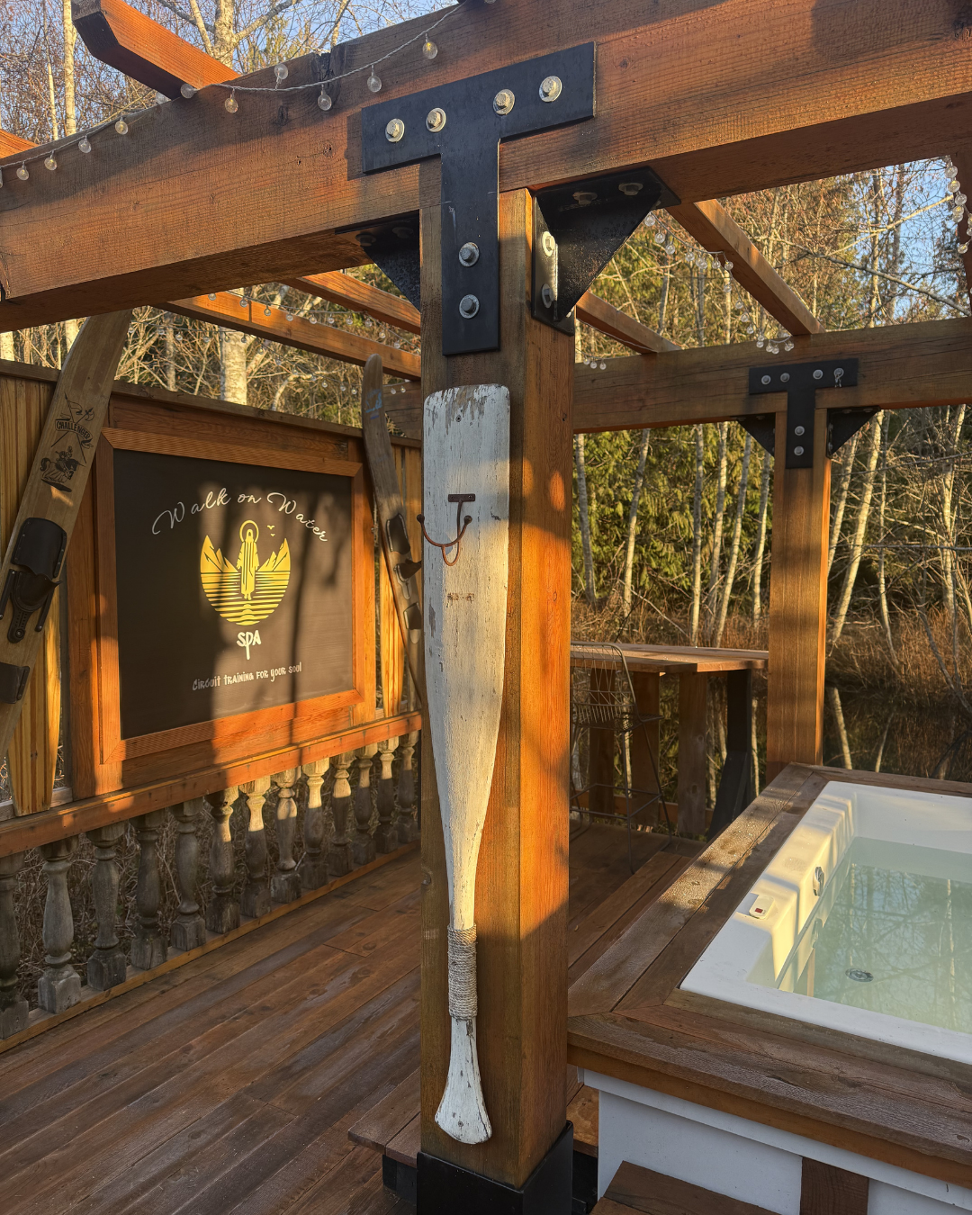Wooden outdoor spa deck with a hot tub, framed by wooden beams with string lights, and decorated with vintage skis and a paddle hanging on a post, beside a sign that says 'Walk on Water' and 'Circuit Training for Your Soul'.