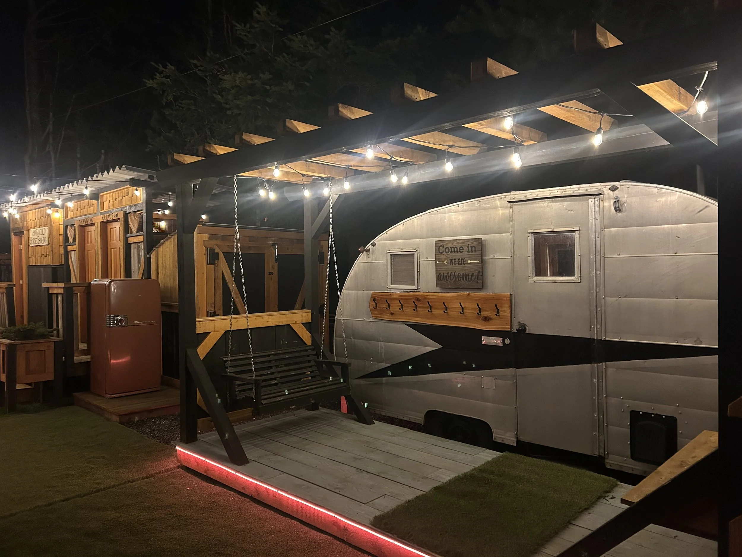 Nighttime scene featuring a shiny silver trailer with a sign that reads 'Come in, we are awesome!' parked on a wooden platform. A wooden swing with chains is hanging from a black pergola with string lights. The setting appears to be an outdoor area w
