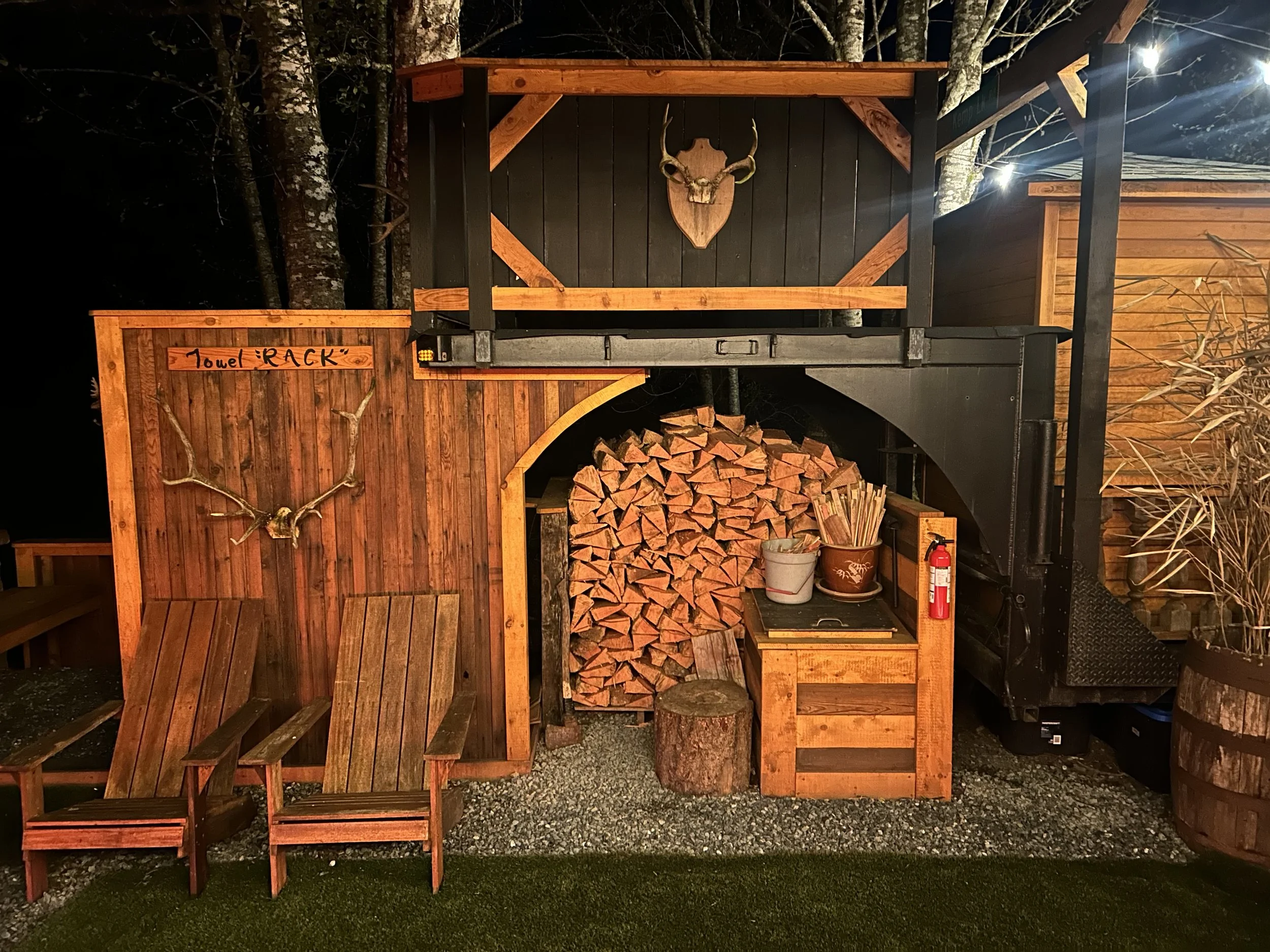 A rustic outdoor wood shelter with a deer skull and antlers mounted on a black and wood-paneled wall. Firewood is stacked underneath a wooden shelf, and a wooden bench is in front. A sign on the left reads 'towel RACK.' A bucket with sticks, a small 