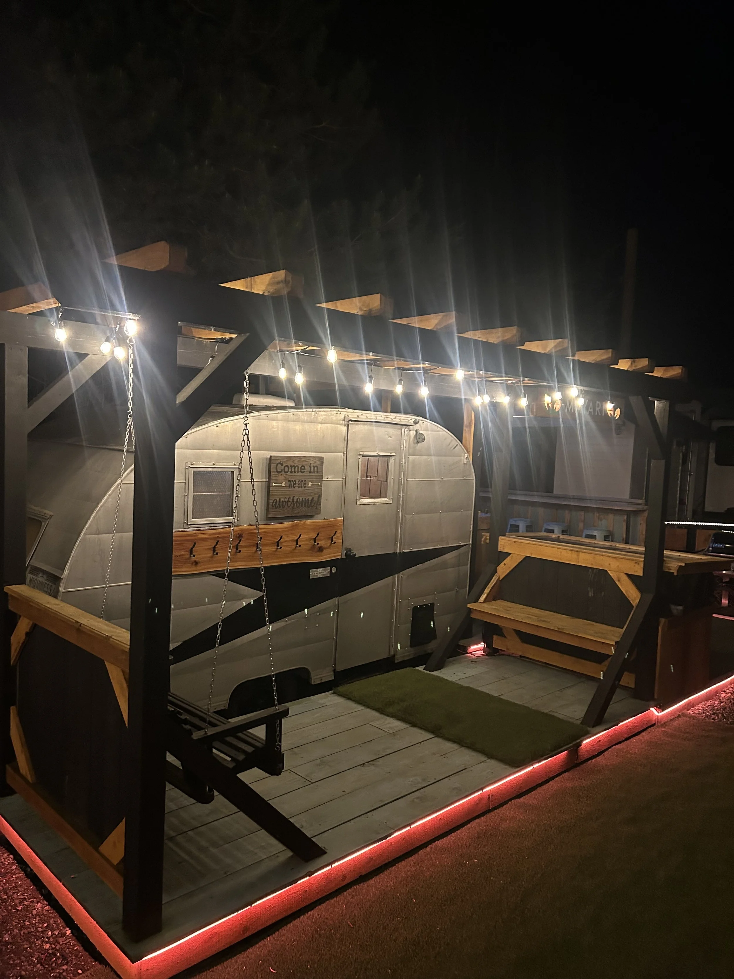 Nighttime outdoor scene featuring a wooden deck with string lights, a metal travel trailer with a sign that says 'Come in we are awesome,' and wooden furniture.