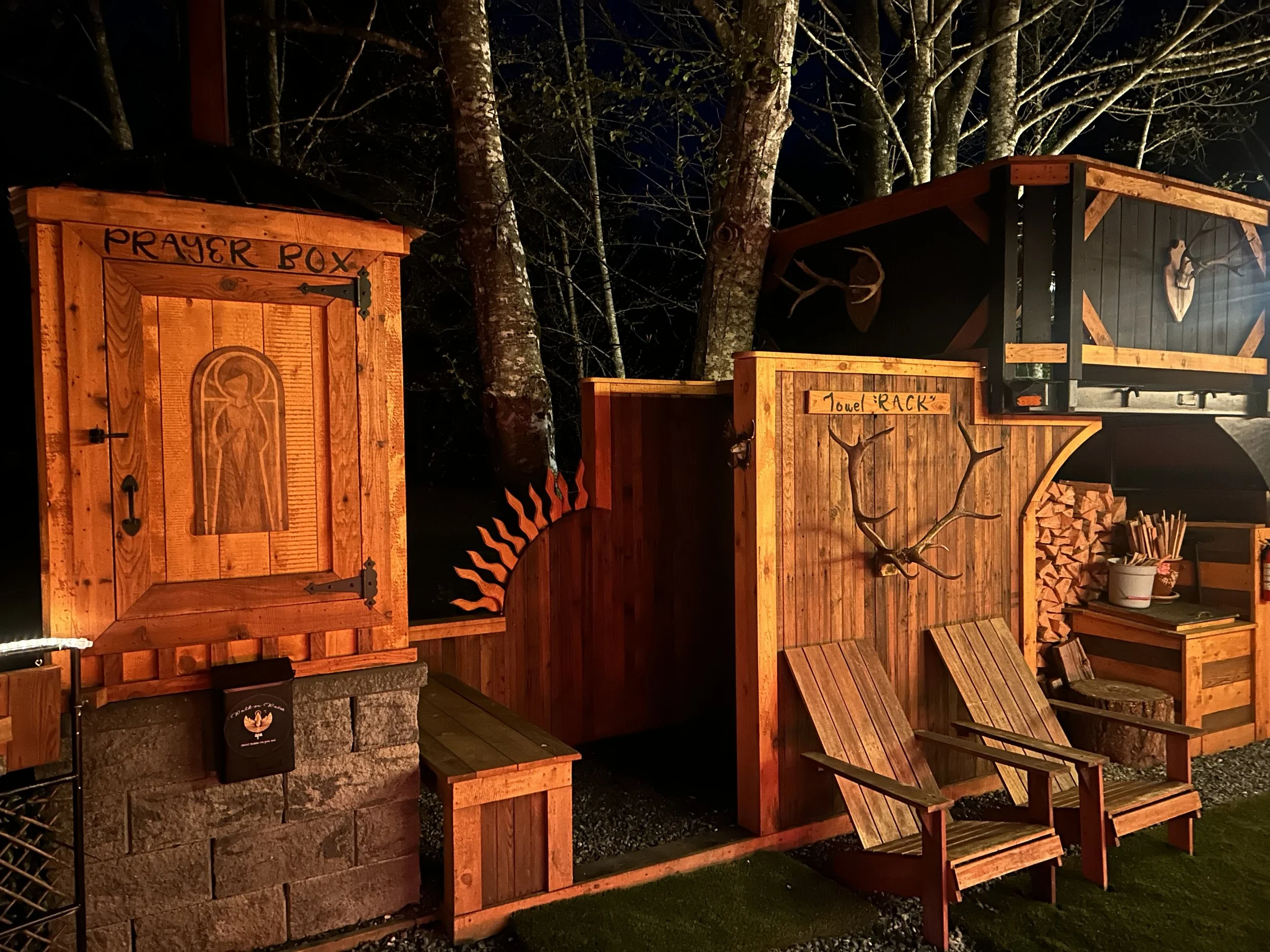 Nighttime outdoor scene with wooden prayer box, towel rack, wooden slats, and mounted deer antlers, trees in background.