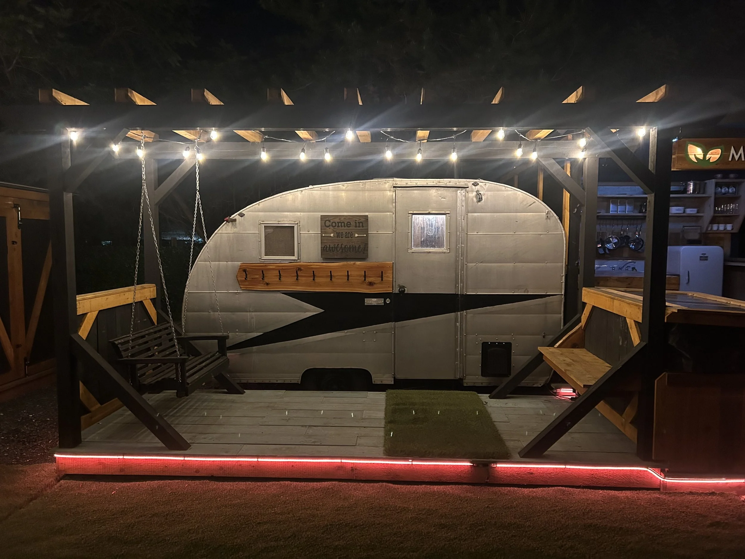 Nighttime outdoor scene featuring a wooden deck with a swing, surrounded by a pergola with string lights, a silver vintage trailer with a small window and sign that reads 'Come in, we are awesome,' nearby shelves with kitchenware, and a sign with a g