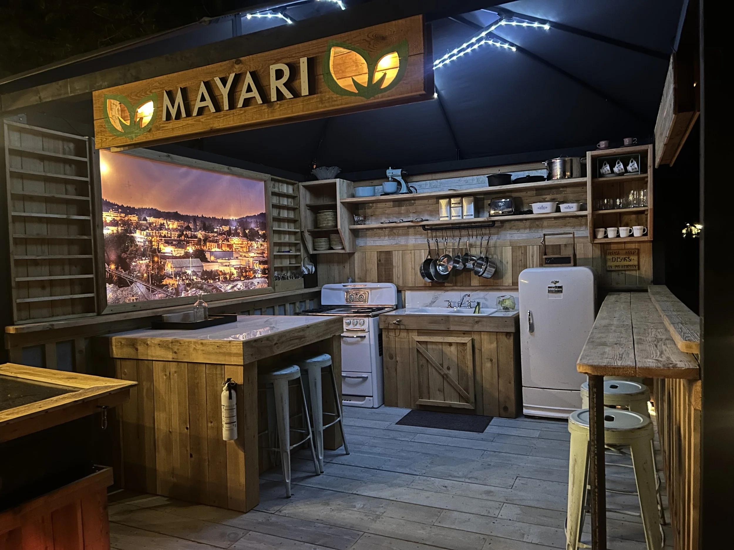 A cozy outdoor kitchen with wooden shelves, cabinets, and countertops, featuring a cooking stove, utensils, and dishes. There is a vibrant cityscape mural on the wall, illuminated by string lights, creating a warm ambience at night.