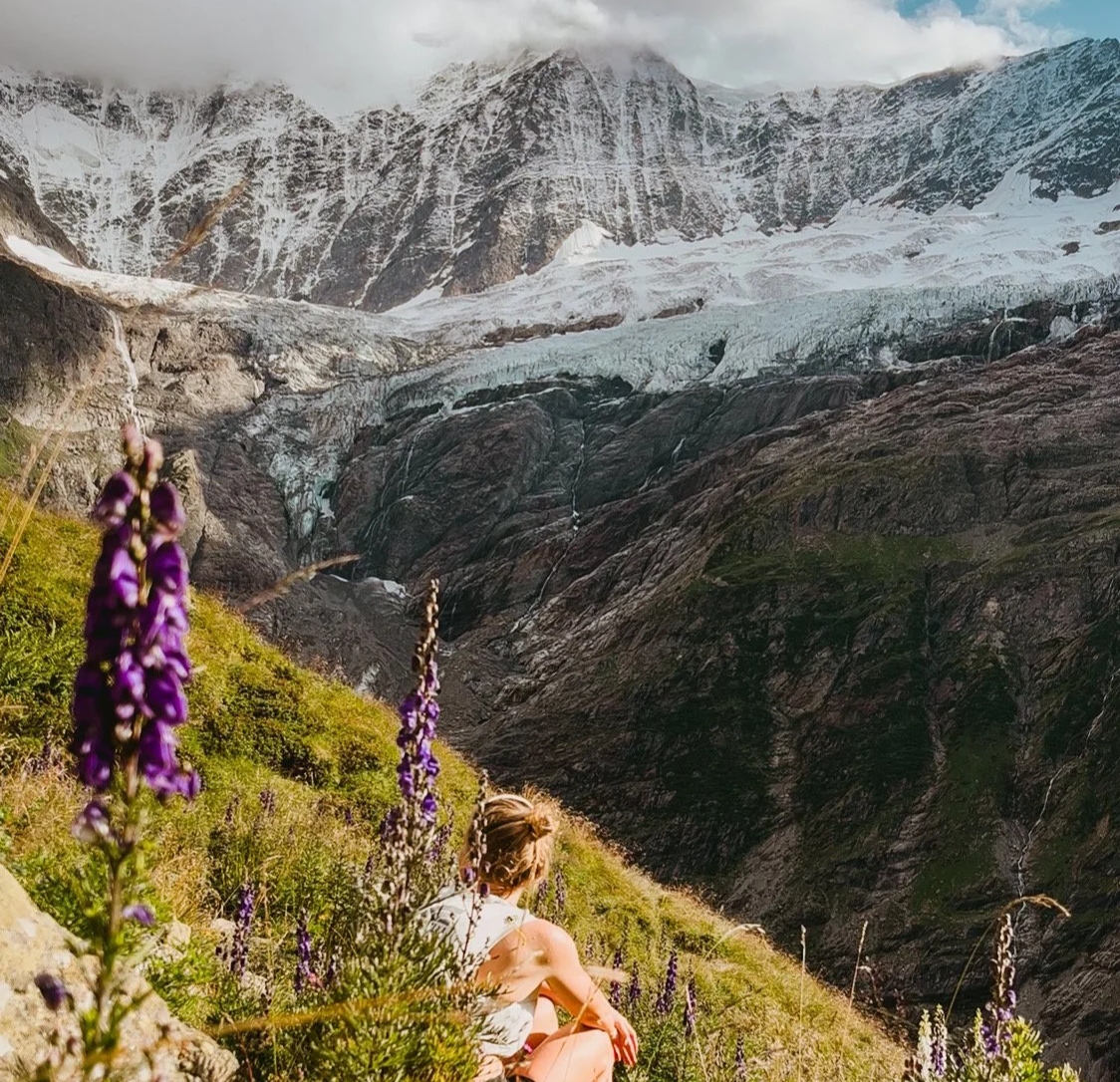 A woman sitting on a grassy hillside with purple flowers, overlooking a mountain landscape with snow-capped peaks, glaciers, and rugged terrain.
