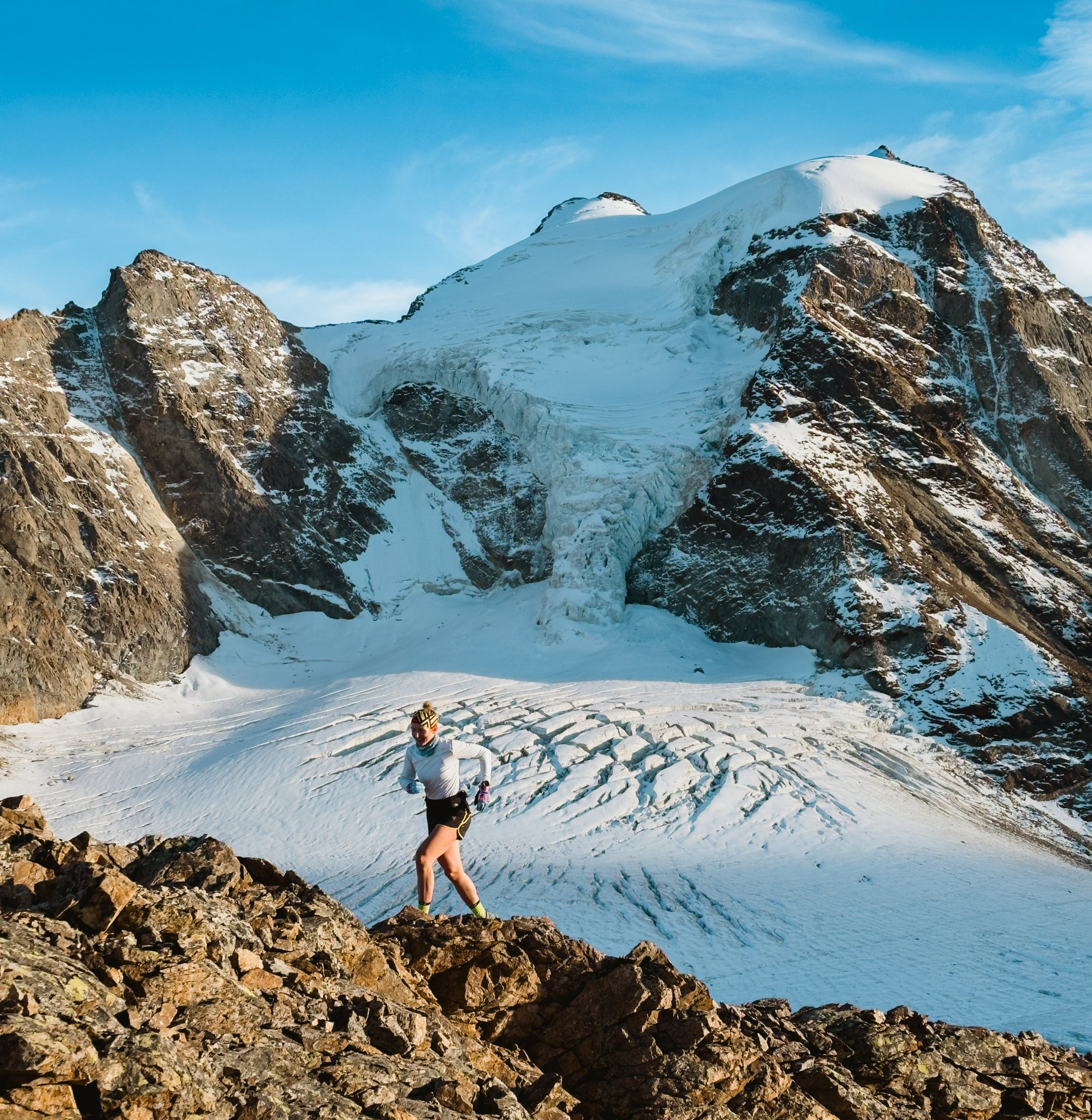 A person running on rocky terrain with a snow-covered mountain and glacier in the background under a partly cloudy sky.