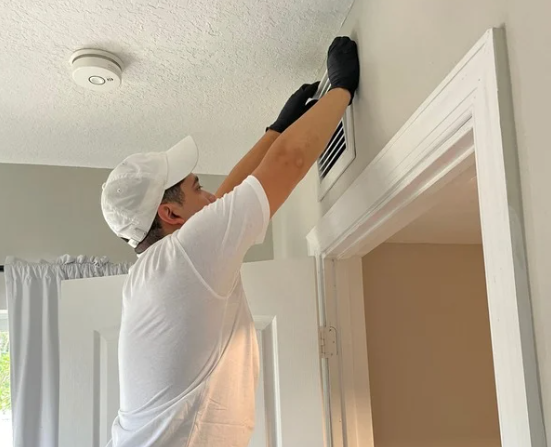 A person wearing a white cap, white t-shirt, and black gloves cleaning an air vent on the ceiling inside a house.