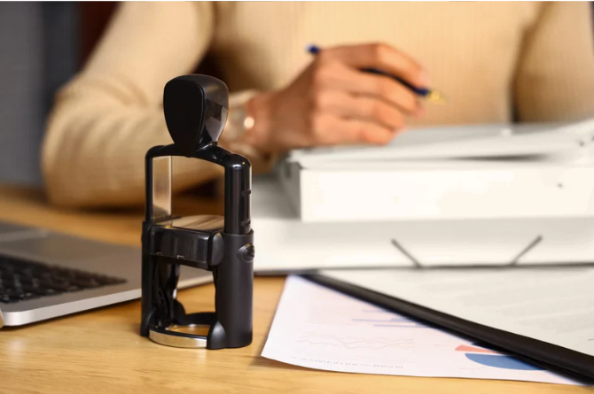 Close-up of a desktop with a hand holding a pen, a document, a laptop, and a black object that appears to be a stamp.