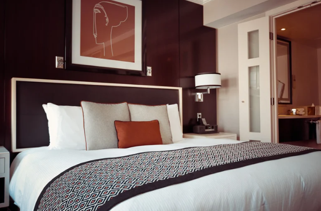 A neatly made hotel bed with white sheets, multiple pillows, and a patterned blanket. Behind the bed, a dark wood headboard extends to a wall with contemporary artwork. A bedside table with a lamp and a phone is visible, and an open doorway reveals p