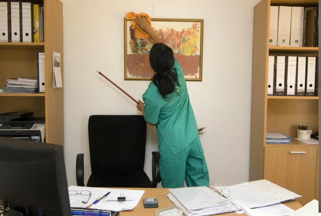 A woman in medical scrubs cleaning a framed picture on an office wall with a cloth, holding a pointing stick in her other hand, in a cluttered office with shelves, a chair, a computer, and various papers.