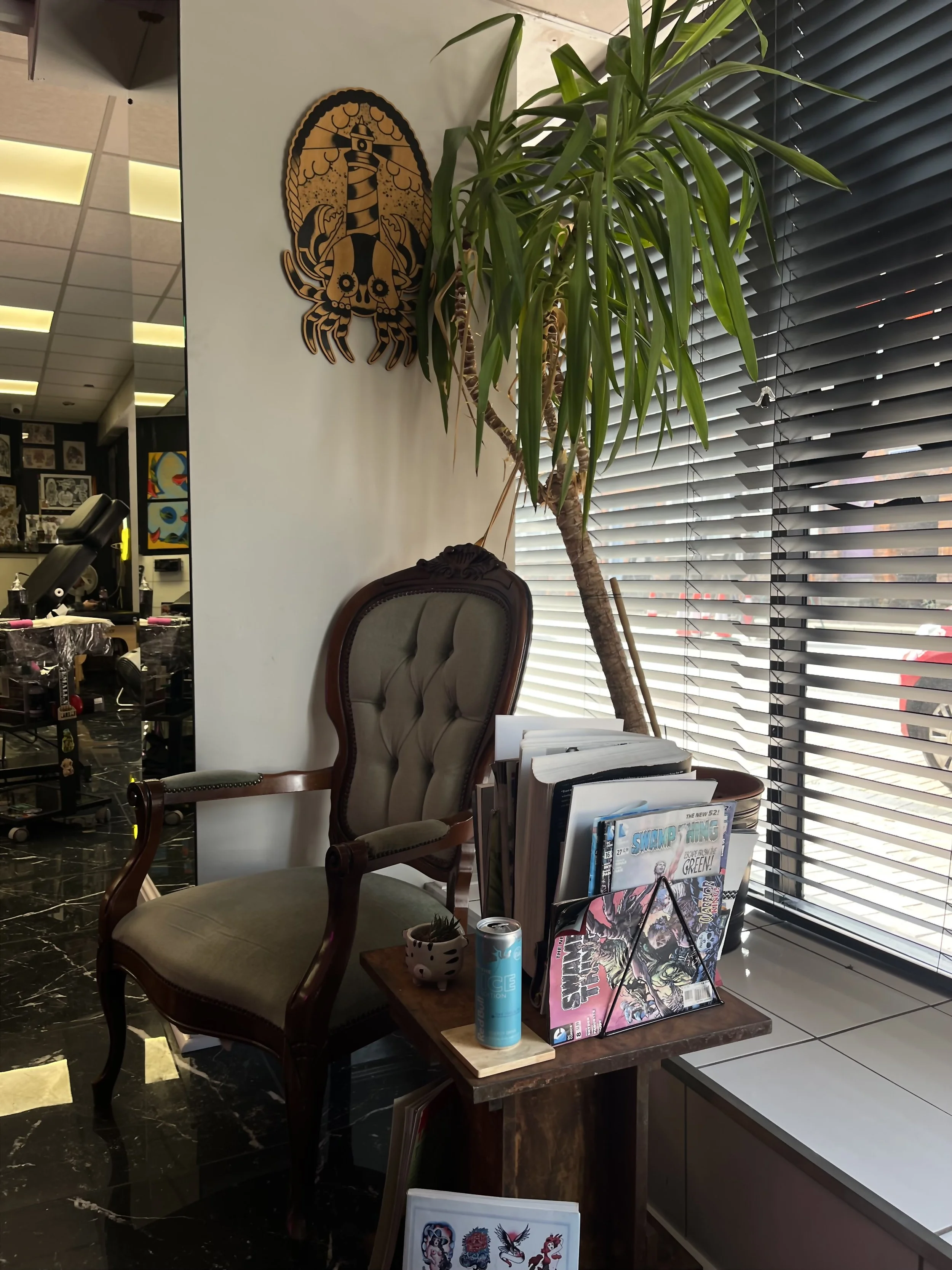 A vintage armchair with wooden frame and tufted upholstery, placed in front of a window with partially closed blinds. Next to the chair, a small wooden table holds a collection of comics, magazines, a can of sparkling water, and a decorative plant. A large indoor plant leans towards the window, with a wall decoration of a lighthouse and sea creatures hanging above