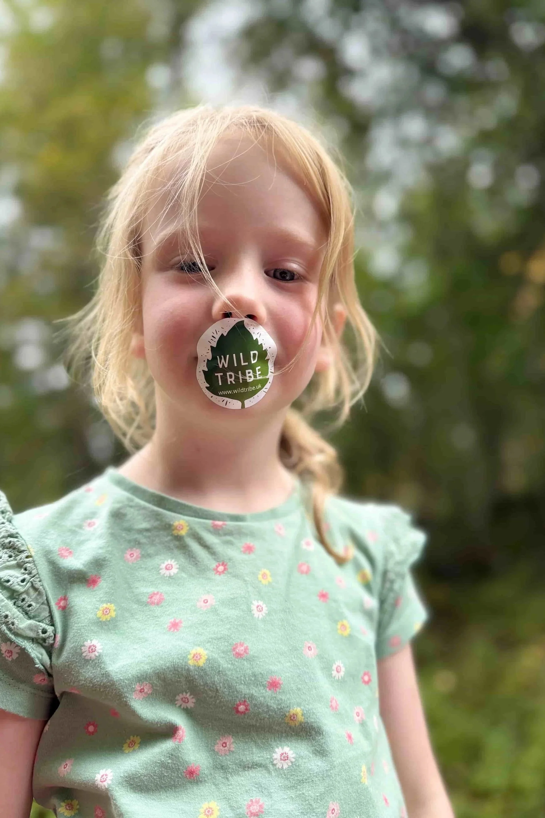 A young girl with blonde hair and a floral green shirt standing outdoors with a blurred green, leafy background, holding a sticker with a tree design in her mouth that reads 'Wild Tribe'