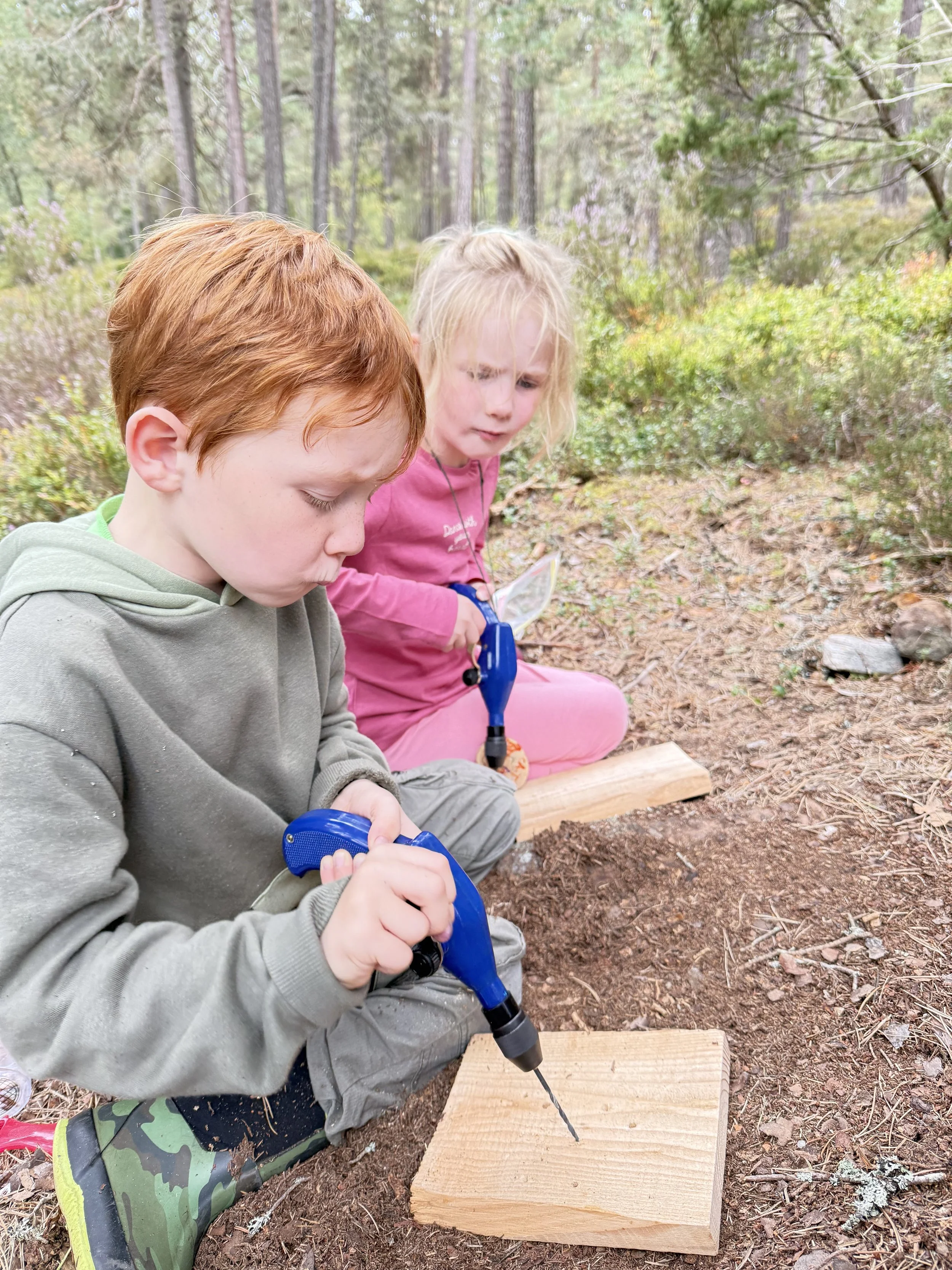 Two children, a boy and a girl, in a forest outdoors, sitting on the ground. The boy is using a blue electric drill on a piece of wood, while the girl watches with curiosity.