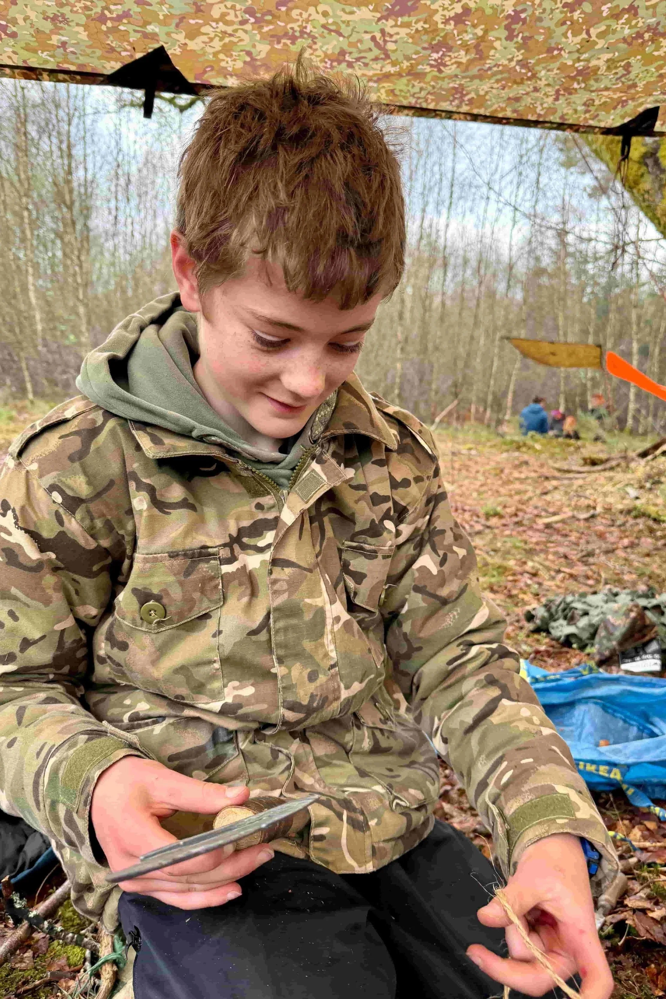 A young boy in camouflage clothing sitting outdoors under a camo canopy, smiling learning bushcraft skills and safe use of tools