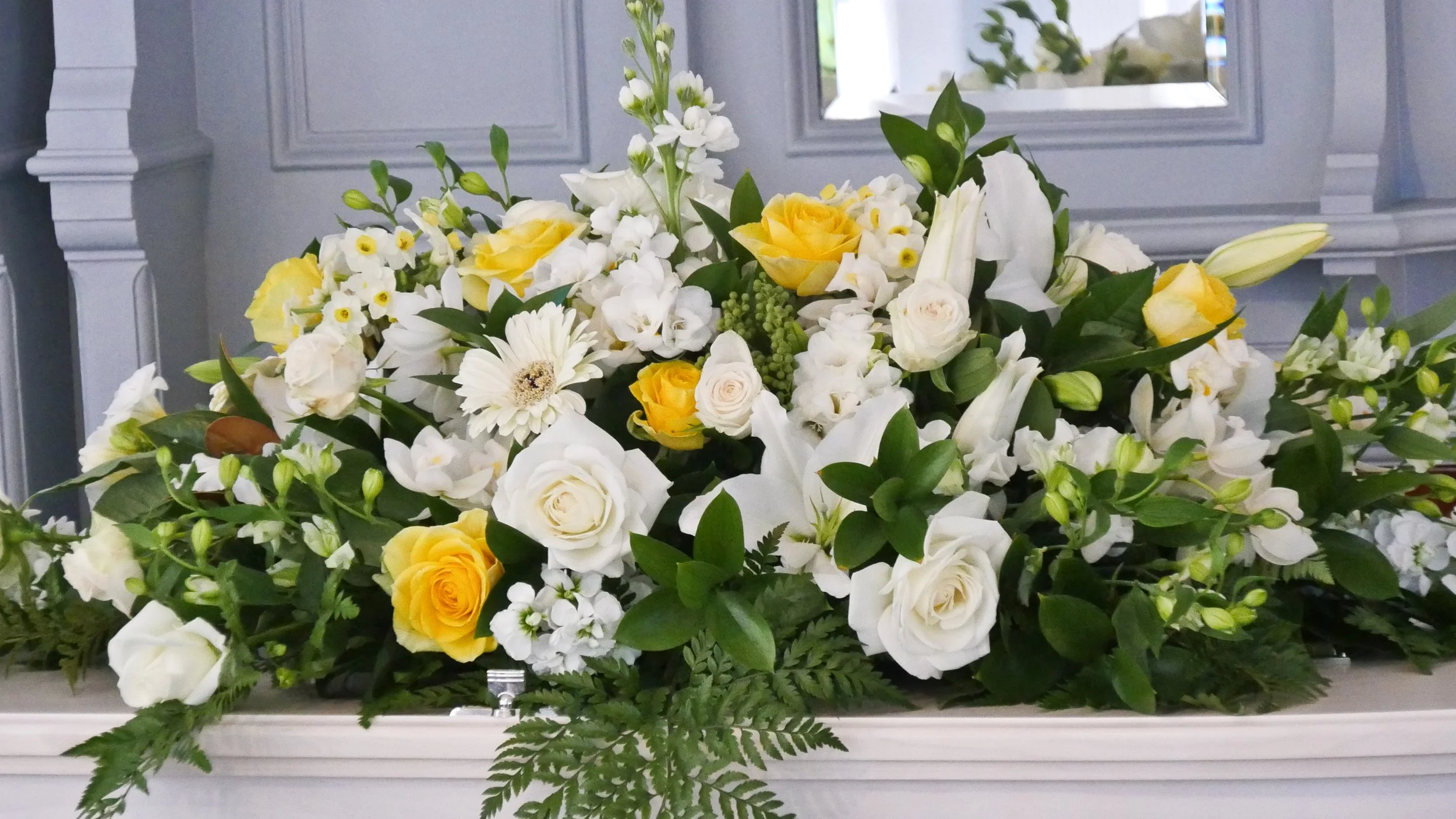 A large casket spray of white and yellow flowers on a white casket surface in front of a gray wall with a mirror.