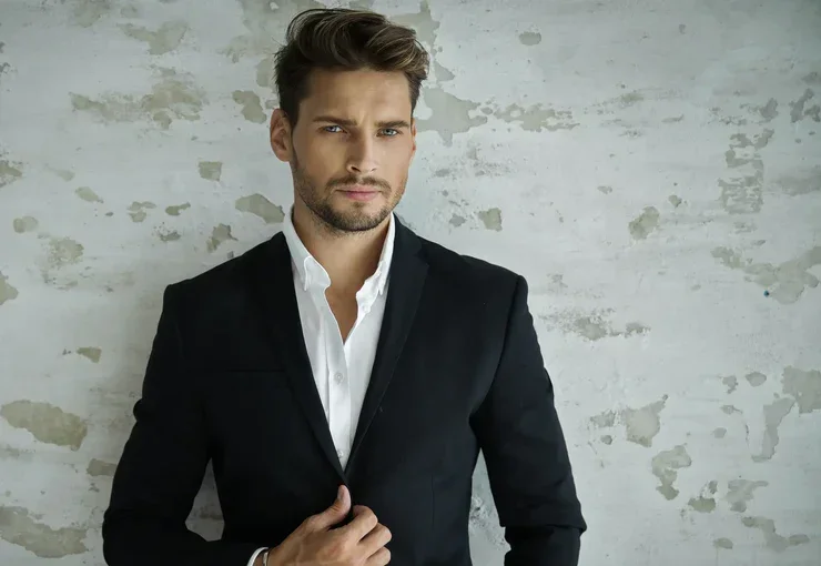 A man in a black suit and white shirt standing against a textured gray wall.