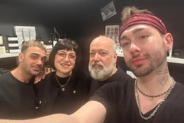 Four people taking a selfie in a kitchen or workroom, smiling and close together. One person is wearing a red bandana and chain, another has glasses and jewelry, and the others have short hair and facial hair.