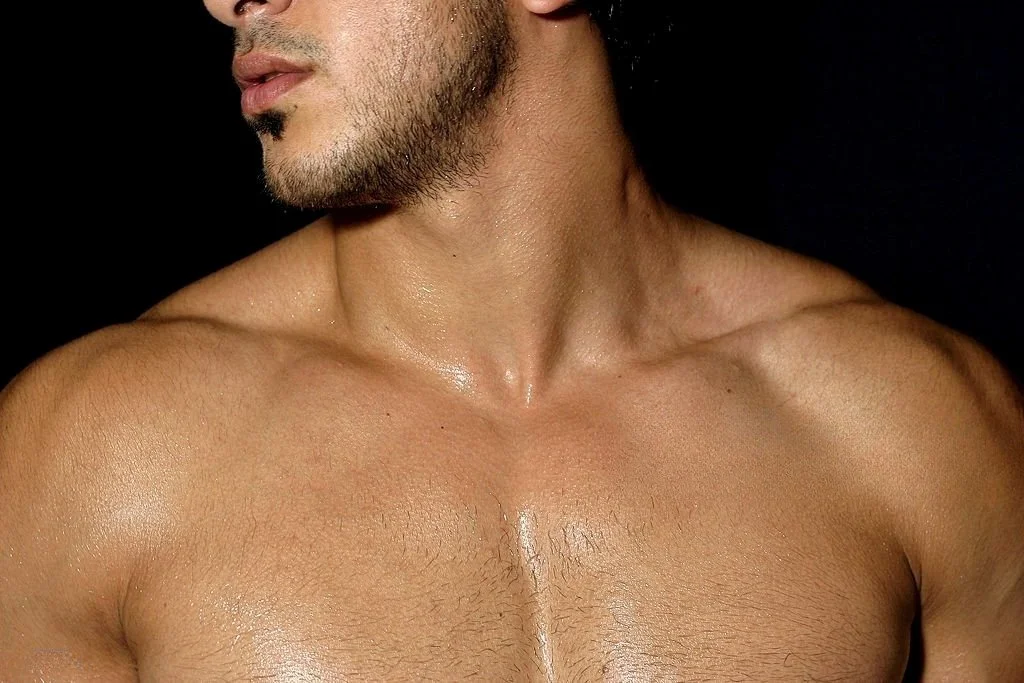 Close-up of a shirtless man with a beard, showing his shoulder, neck, and part of his face against a black background.