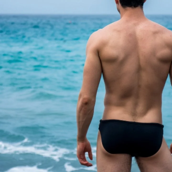 A man standing at the edge of the ocean wearing black swim trunks, viewed from behind, with the sea and sky in the background.