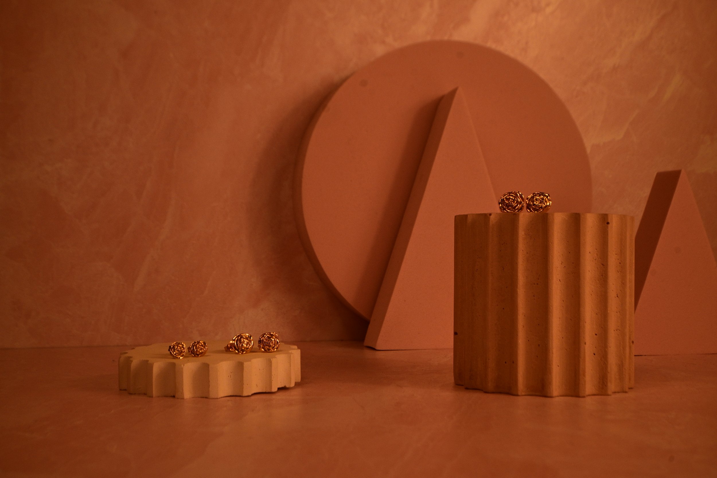 A decorative display of blossoming 18k gold rose stud earrings in three scaling sizes.  Earrings are displayed on a stylish cylindrical pedestal with geometric shapes beautifying the background.