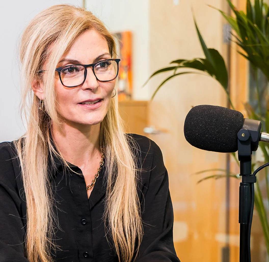 A woman with long blonde hair wearing glasses and a black shirt sitting in front of a microphone during an interview or podcast recording.
