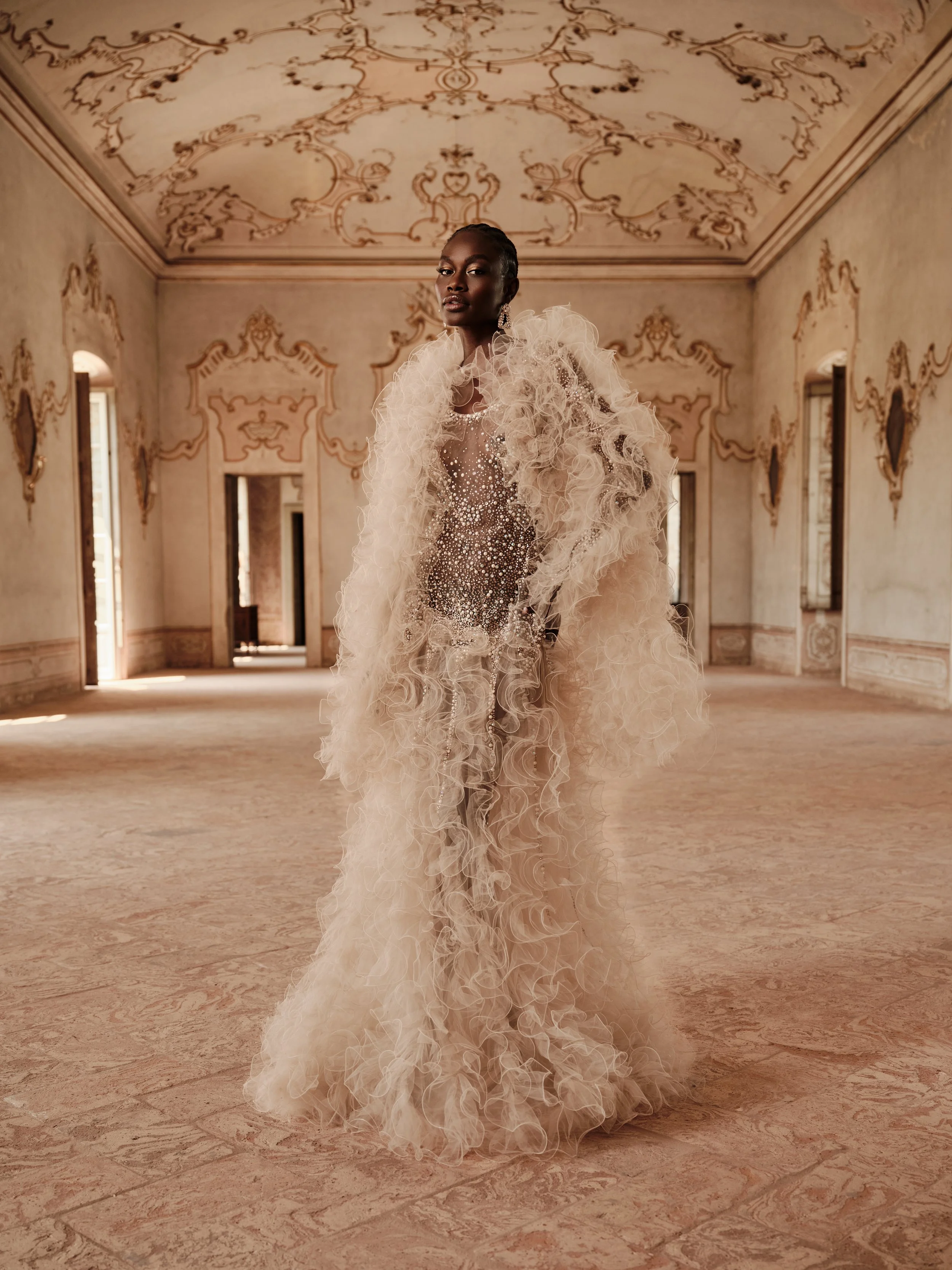 A woman in an ornate, ruffled gown stands in a grand, classical-style room with decorated walls and high ceilings.