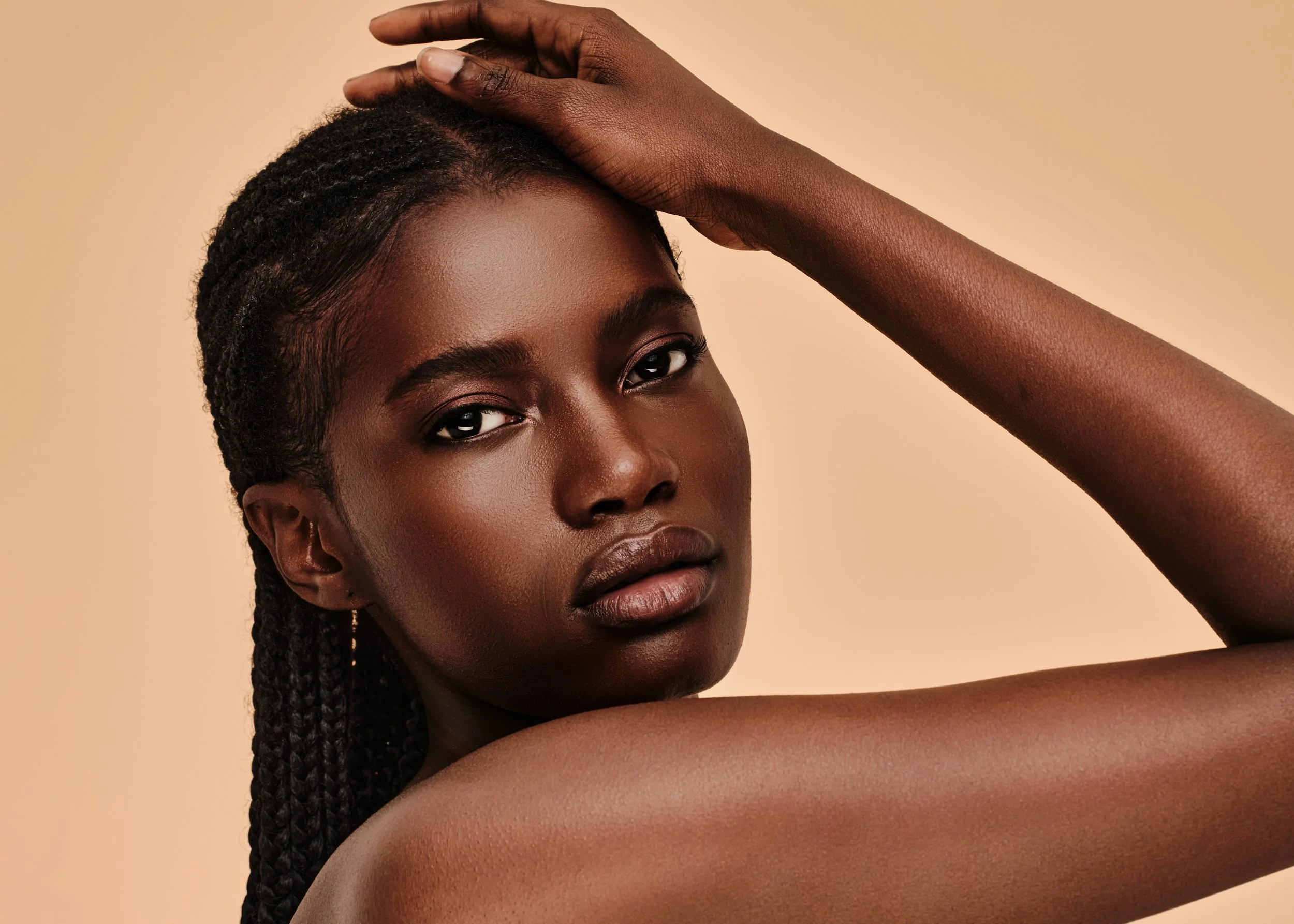 Close-up portrait of a young woman with dark skin, braided hair, and natural makeup, posing with her arm across her forehead against a beige background.