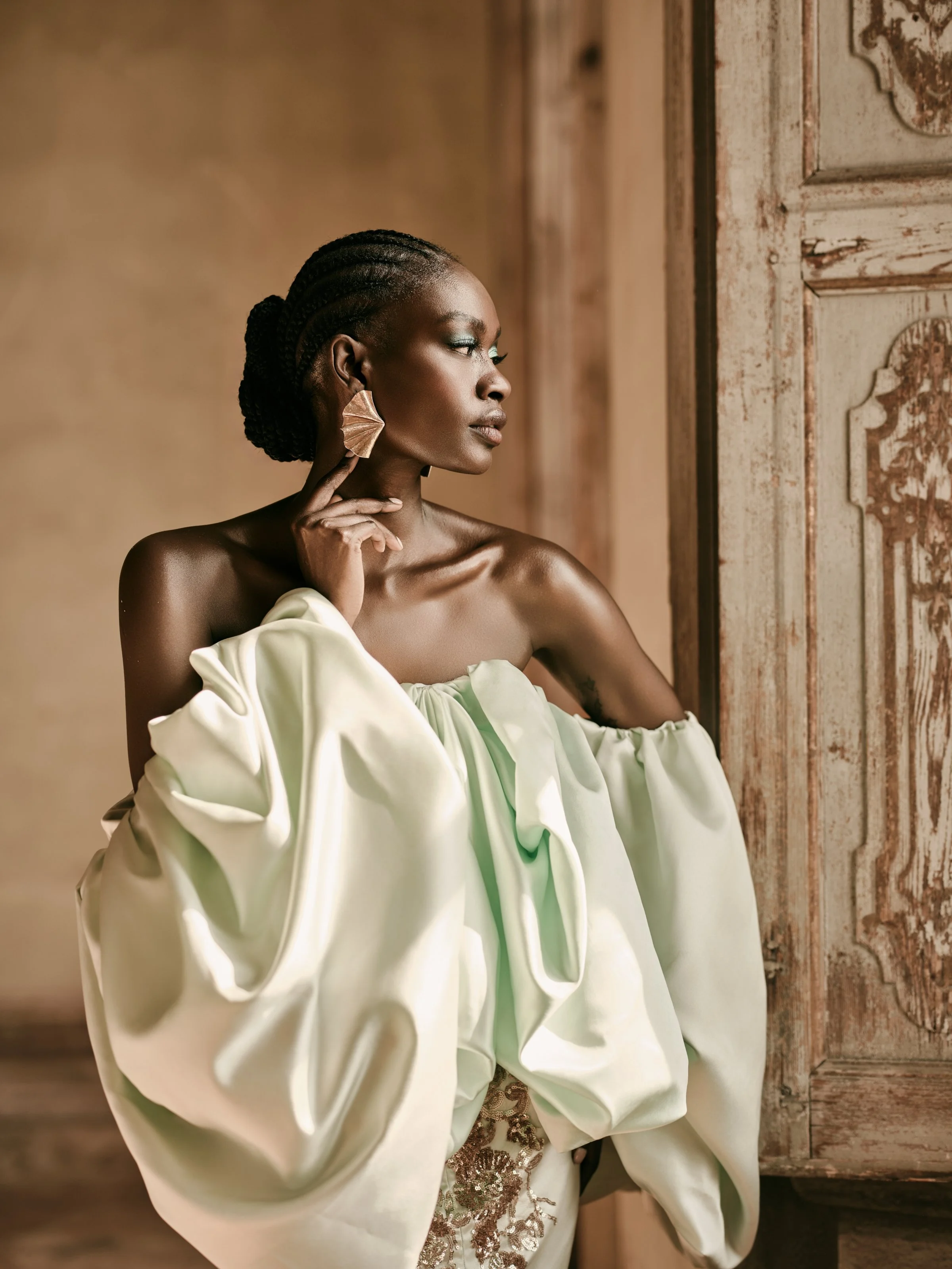 A woman with dark skin and braided hair, wearing a large off-shoulder white gown and statement earrings, is posing indoors near a vintage wooden door and textured wall, looking to the side.