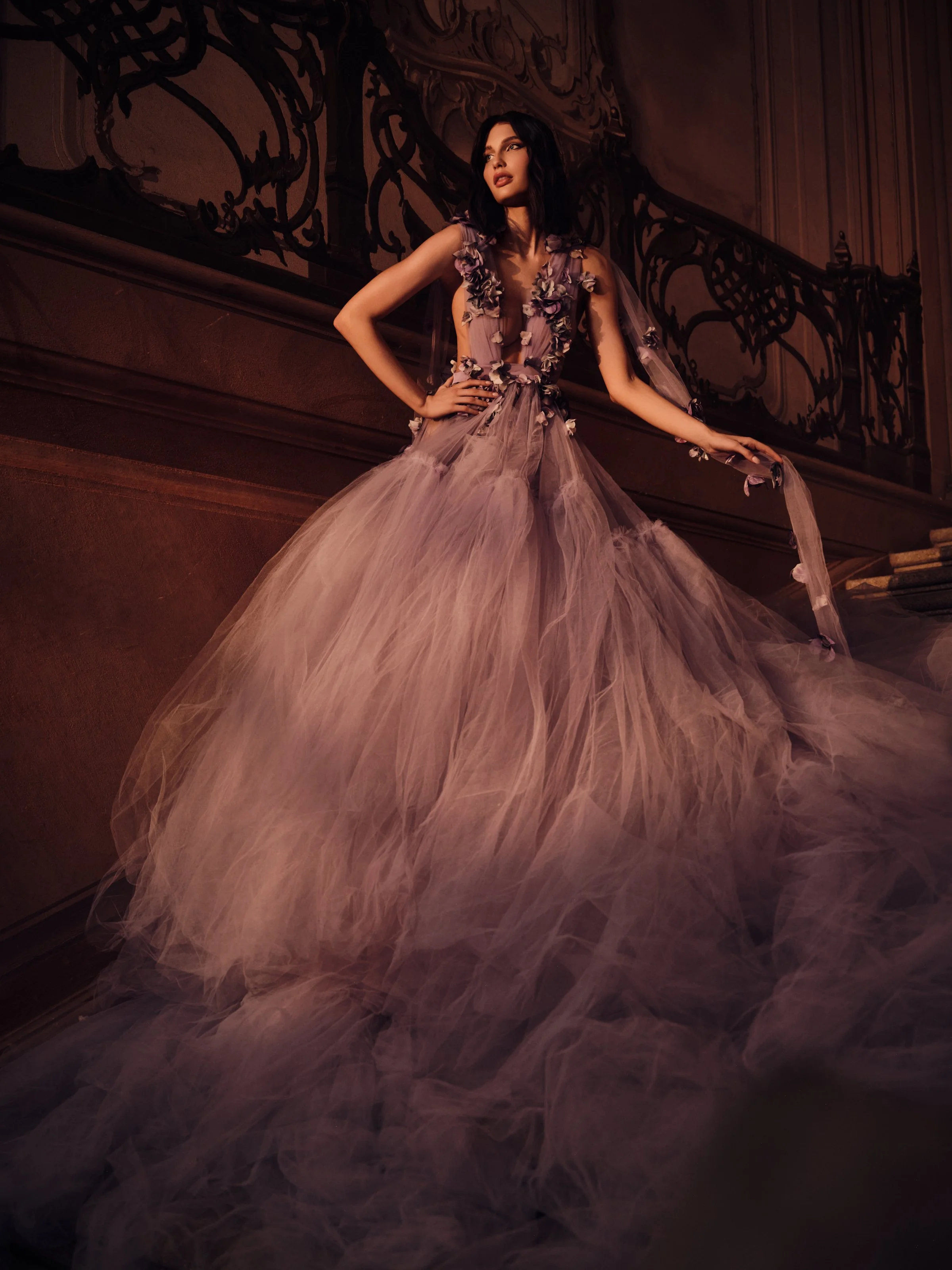 A woman in a flowing, purple and pink tulle gown with floral embellishments standing on a staircase with ornate railing in a dimly lit, elegant interior.